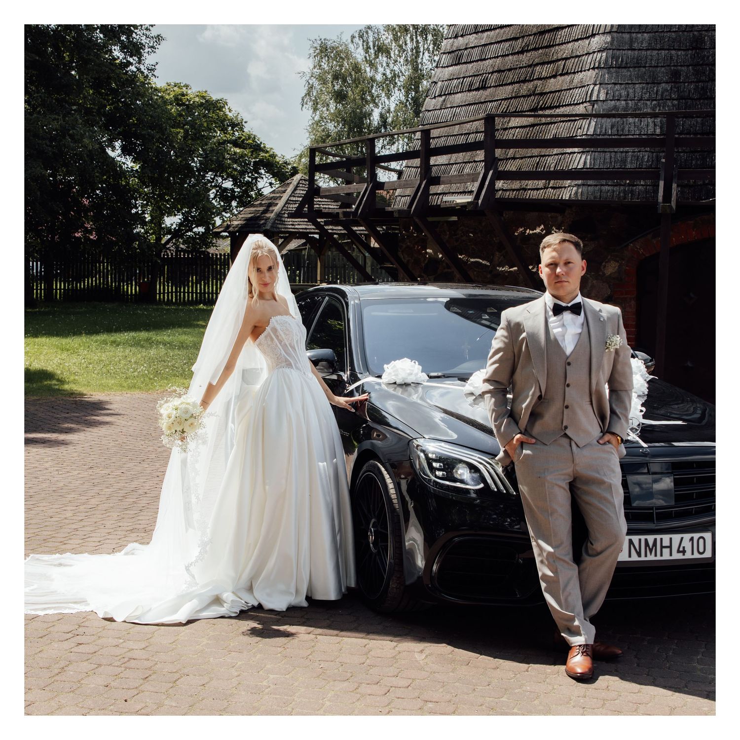 Bride in white gown and groom in gray suit pose beside a black luxury car decorated with white ribbons.