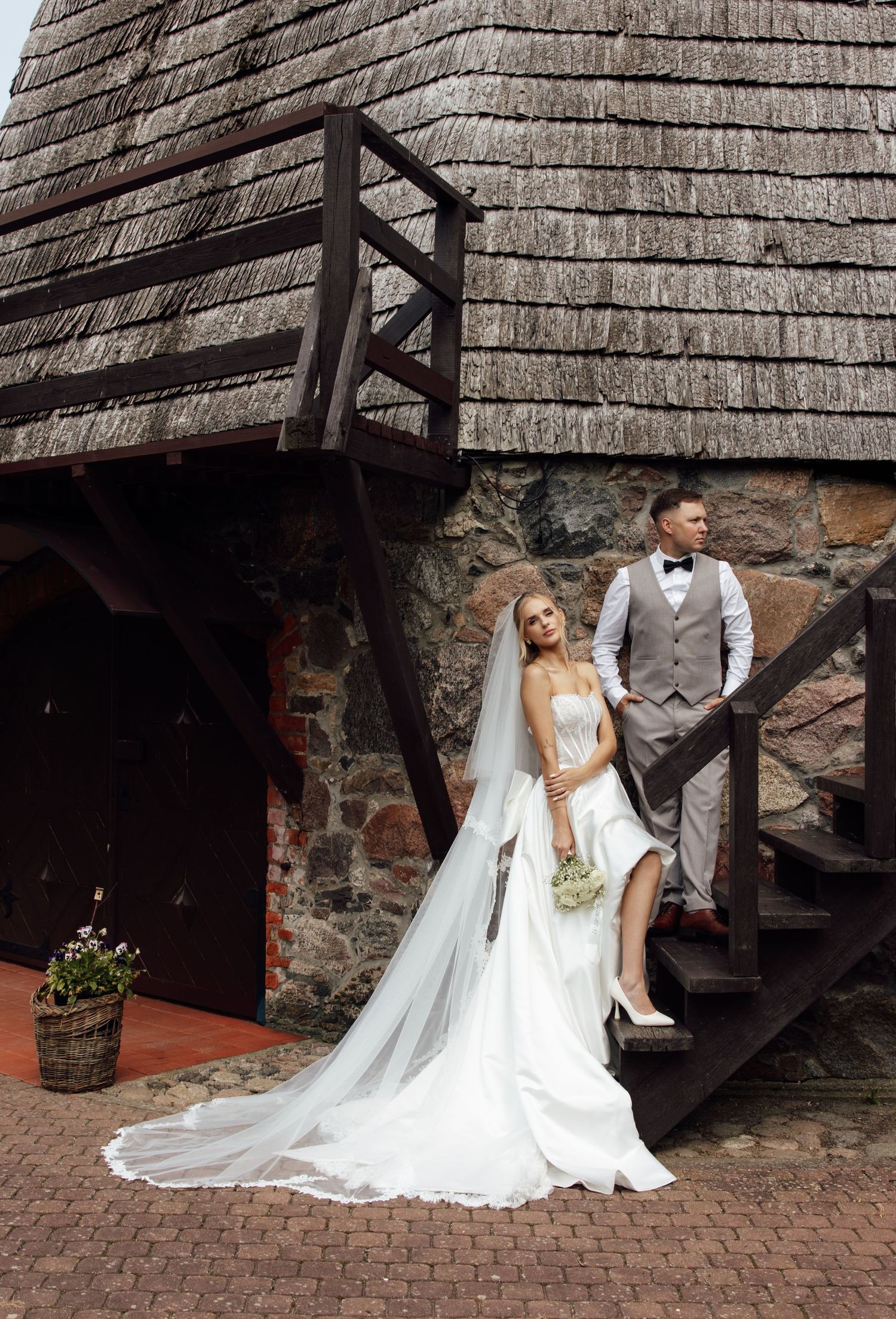 Elegant bride and groom pose on wooden stairs beside a stone wall and thatched roof building.