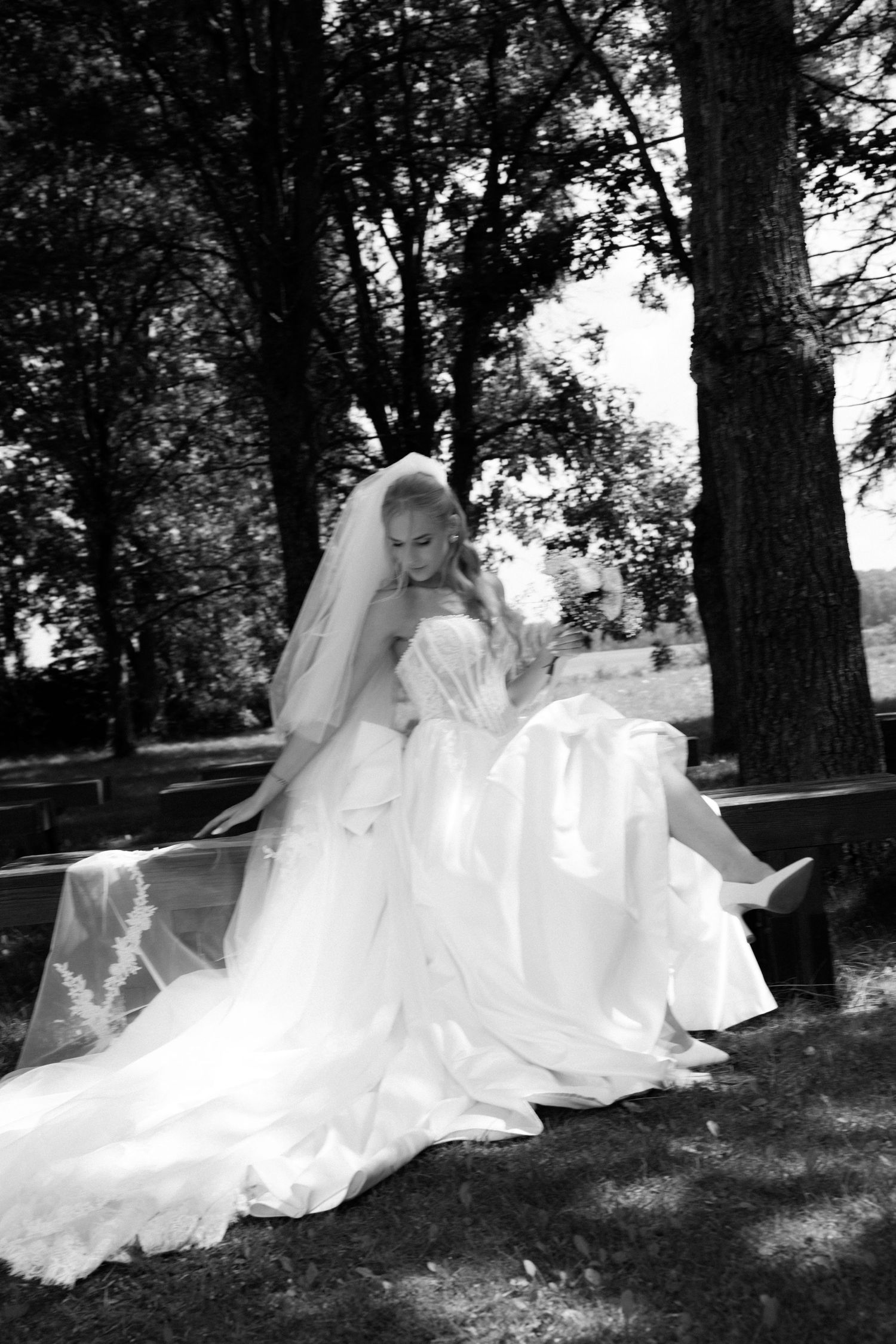 Black and white photo of a bride in a flowing white gown and veil seated on a bench surrounded by trees.