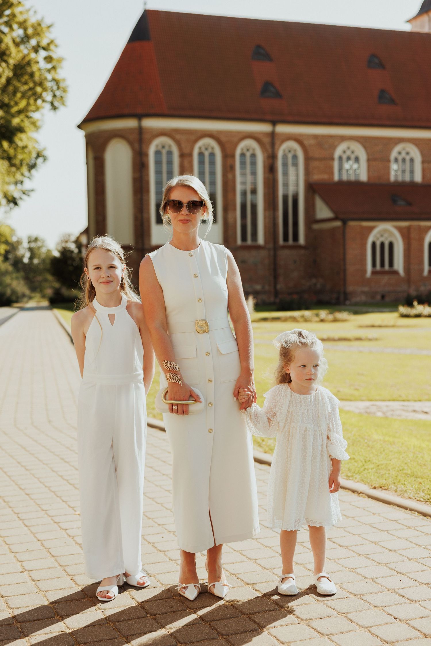 Small children dressed in all white stand together on a stone pathway near a traditional brick church.