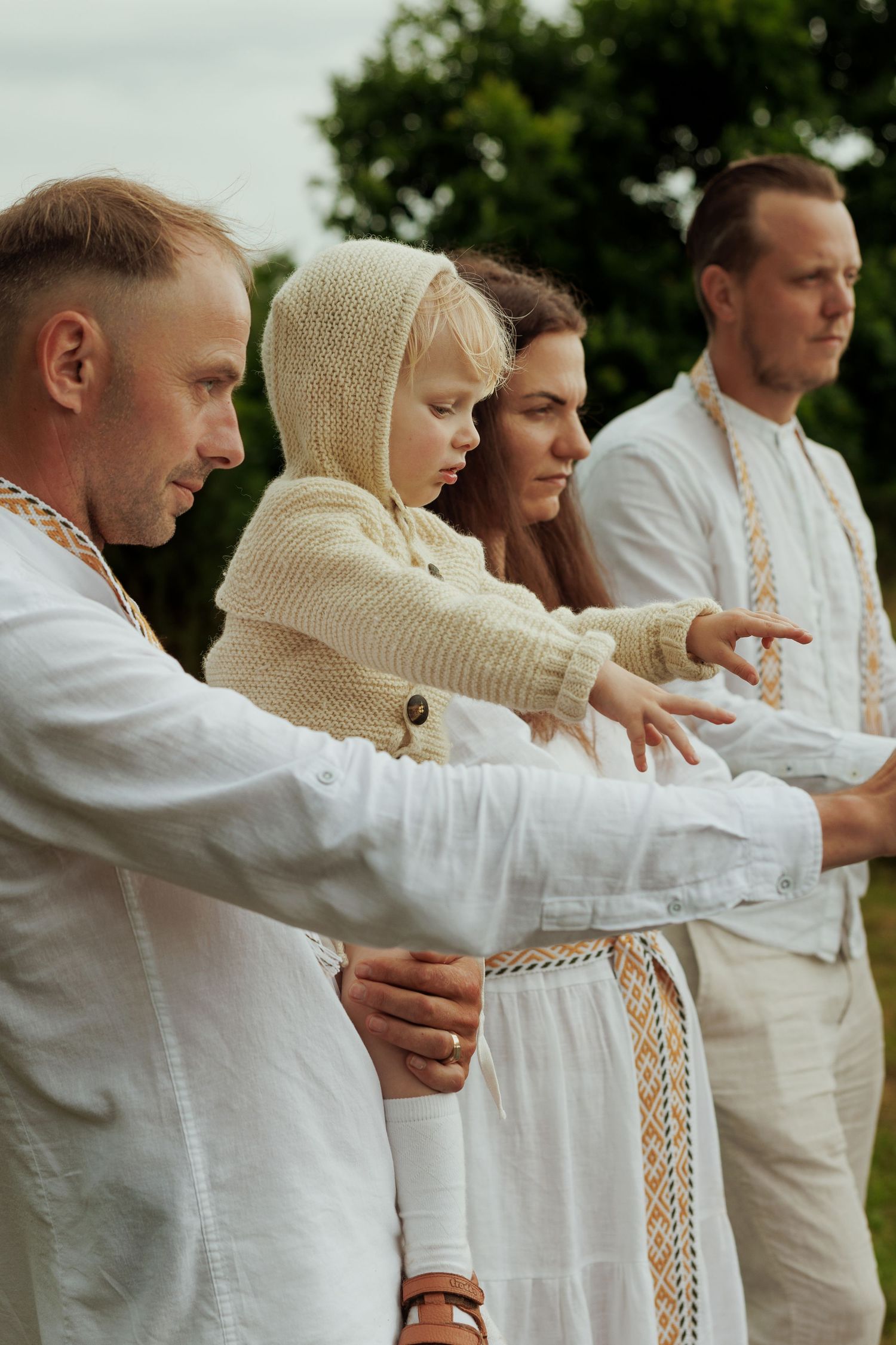 Family in white embroidered folk attire pointing outdoors, including a child in a knit bonnet.