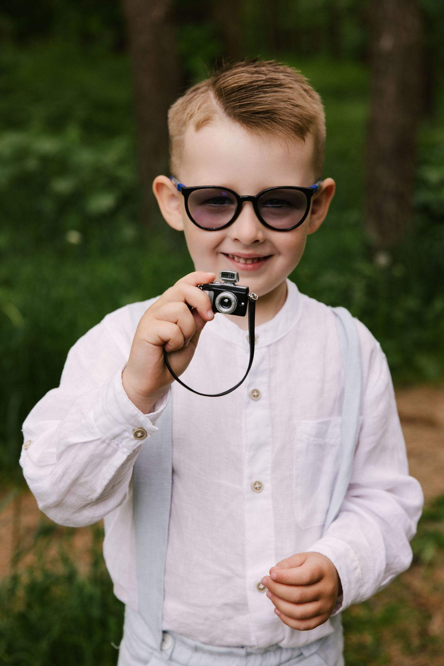 Child in white shirt and glasses playing with vintage camera in wooded setting.