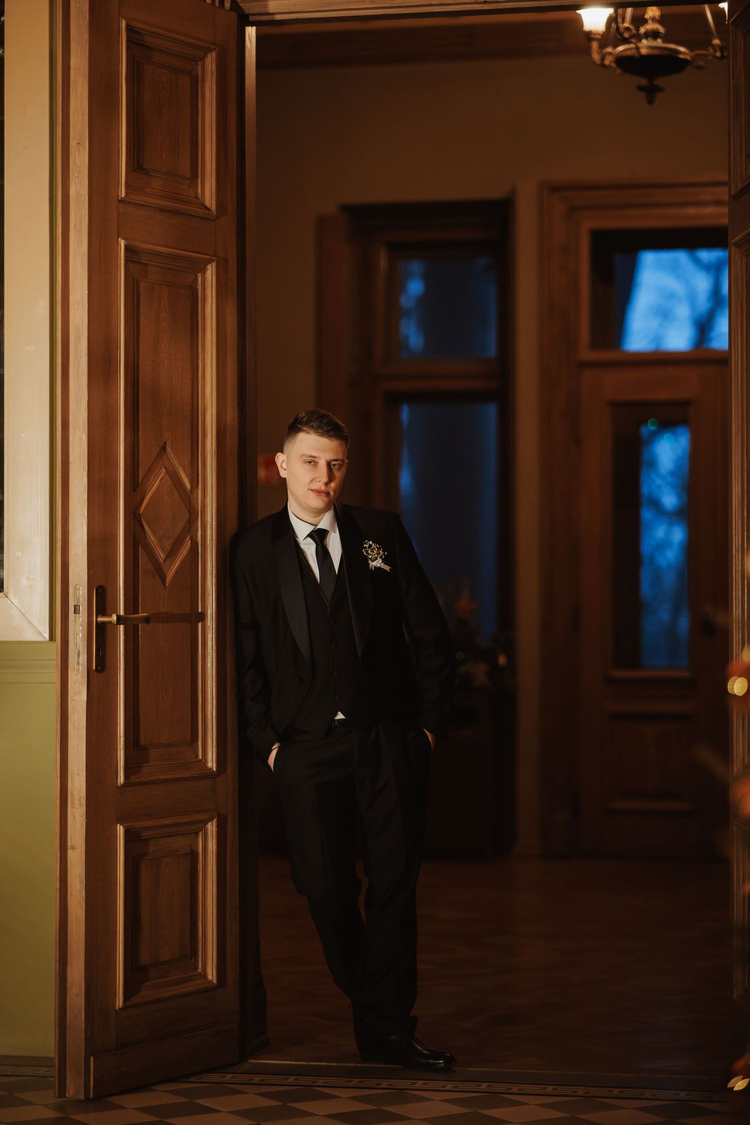 Elegant wedding couple poses in a dimly lit hallway with ornate wooden doors and warm lighting.