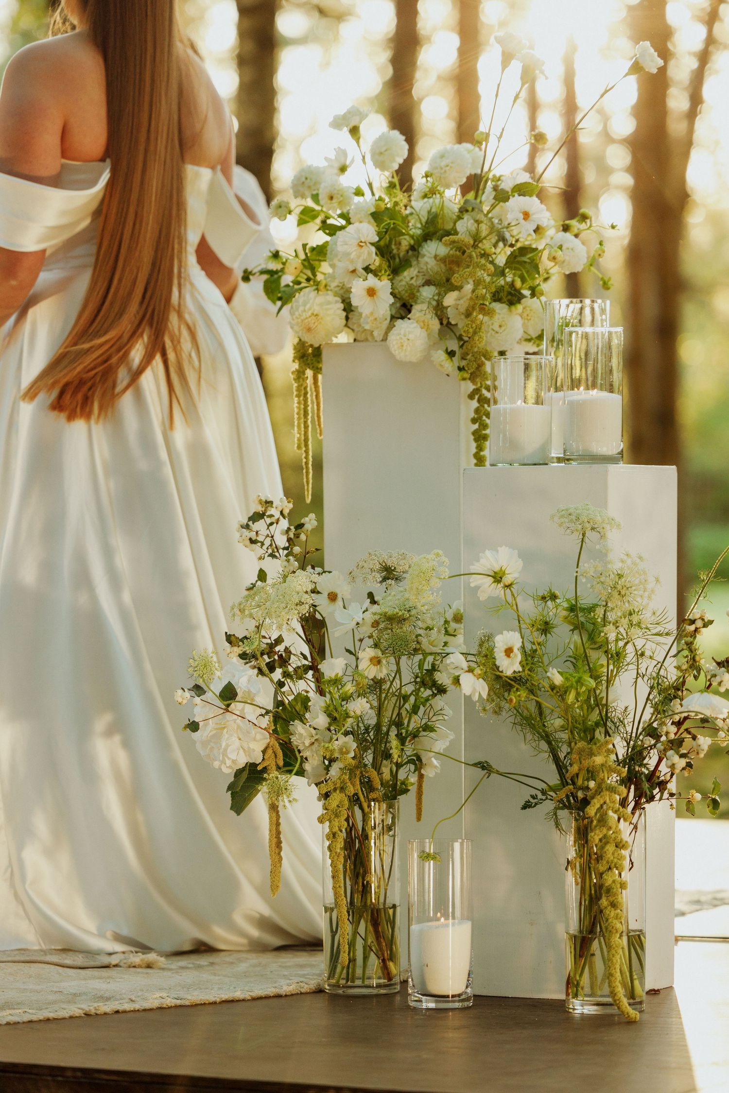 White floral arrangements in glass vases against sunlit forest backdrop at outdoor wedding.
