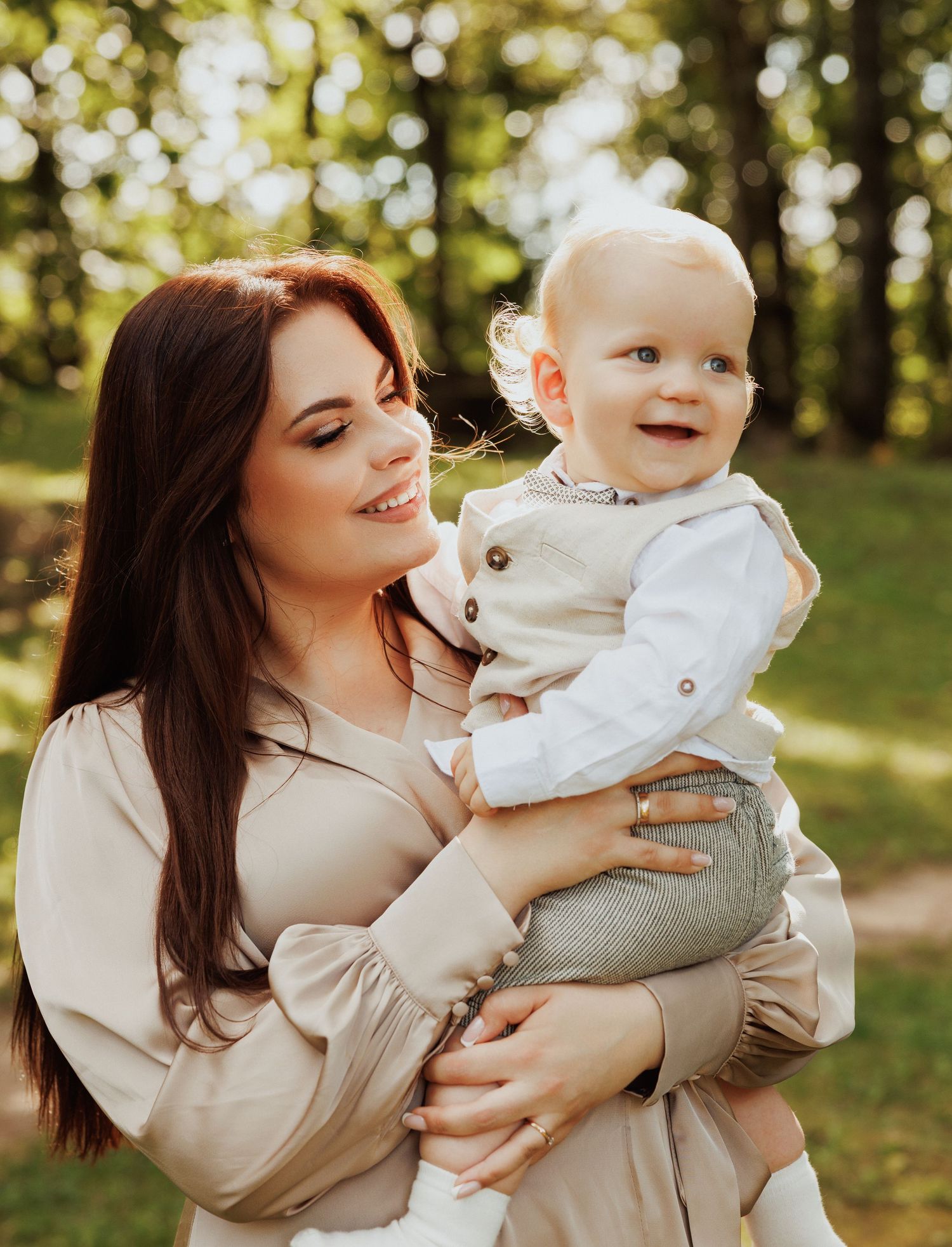 Mother and baby in coordinating beige outfits pose outdoors in sunlit forest setting.