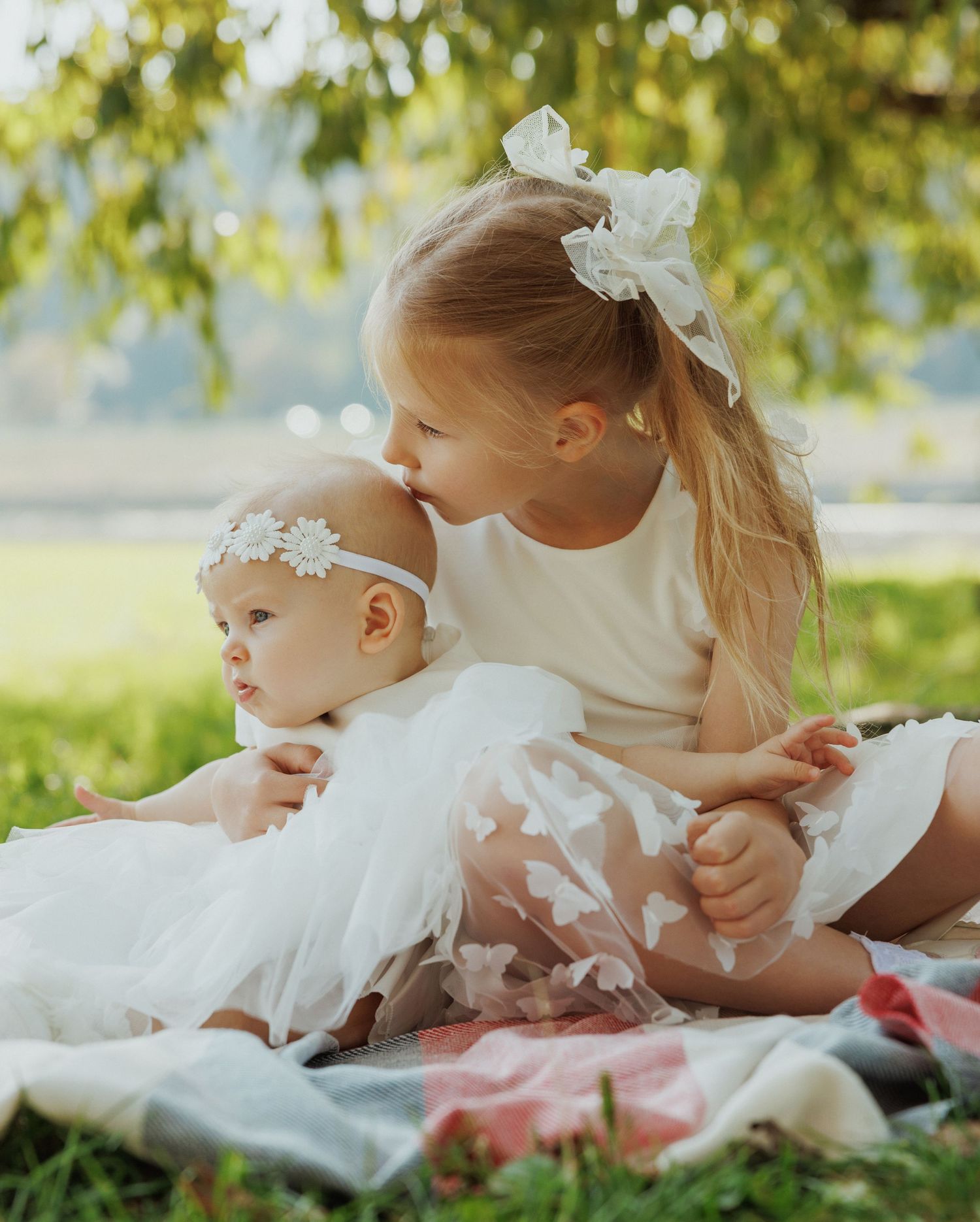 Two children in white dresses sit together on a blanket outdoors with sunlit trees in the background.