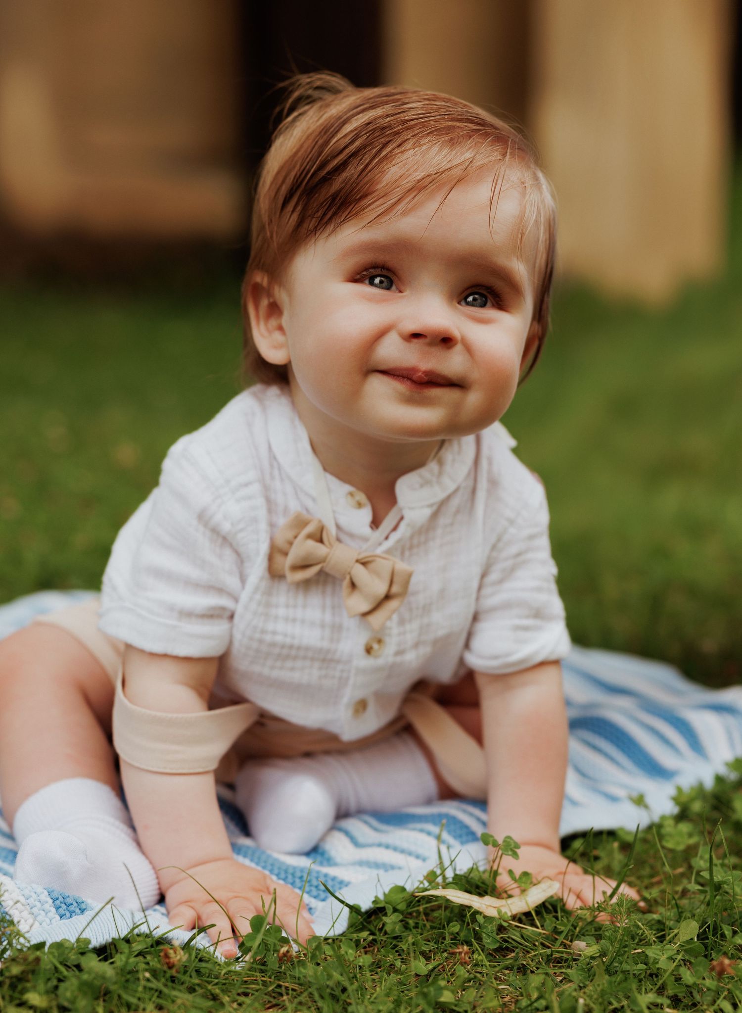 Baby in white outfit lies on blue blanket in grass while crawling and smiling during outdoor photo session.