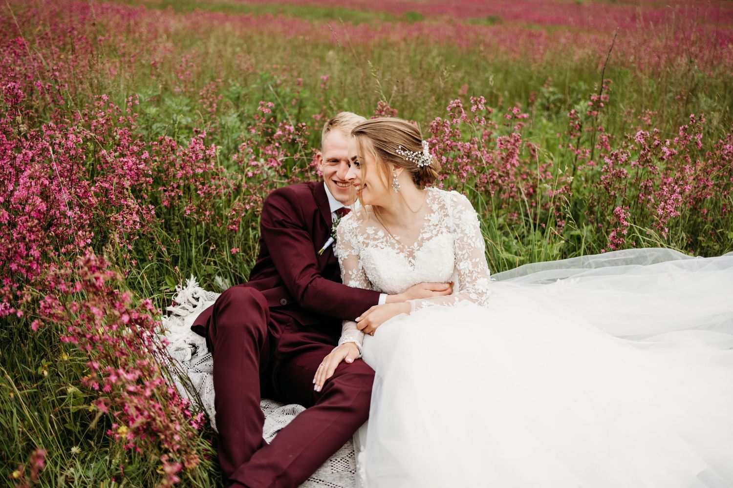 A romantic wedding photo in a field of vibrant pink wildflowers with flowing white dress and burgundy suit.