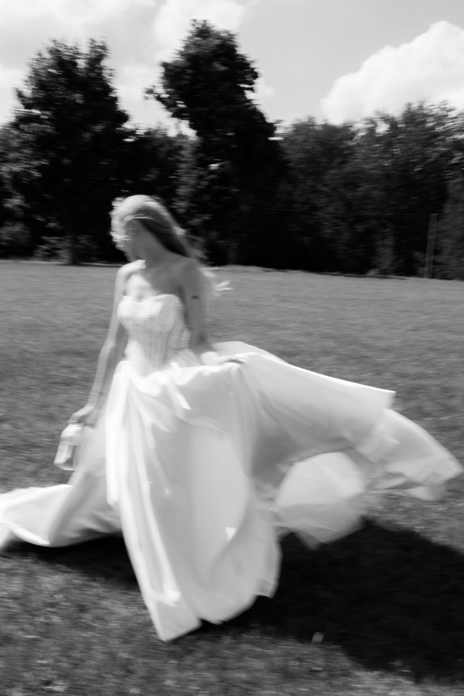 Bride in flowing white gown twirling outdoors in black and white photo with trees in background.