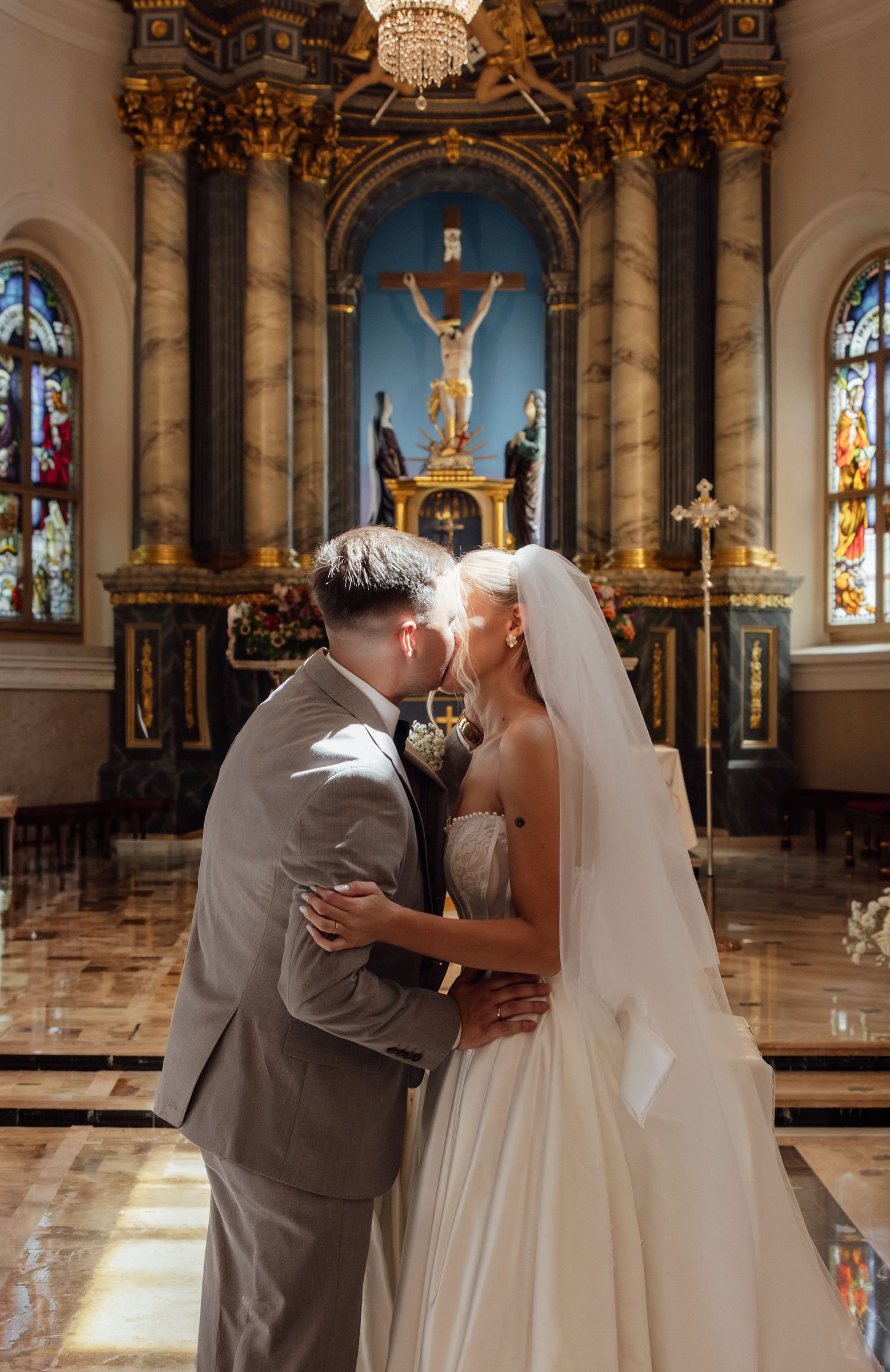 Bride and groom share first kiss at ornate church altar with crucifix and stained glass windows.