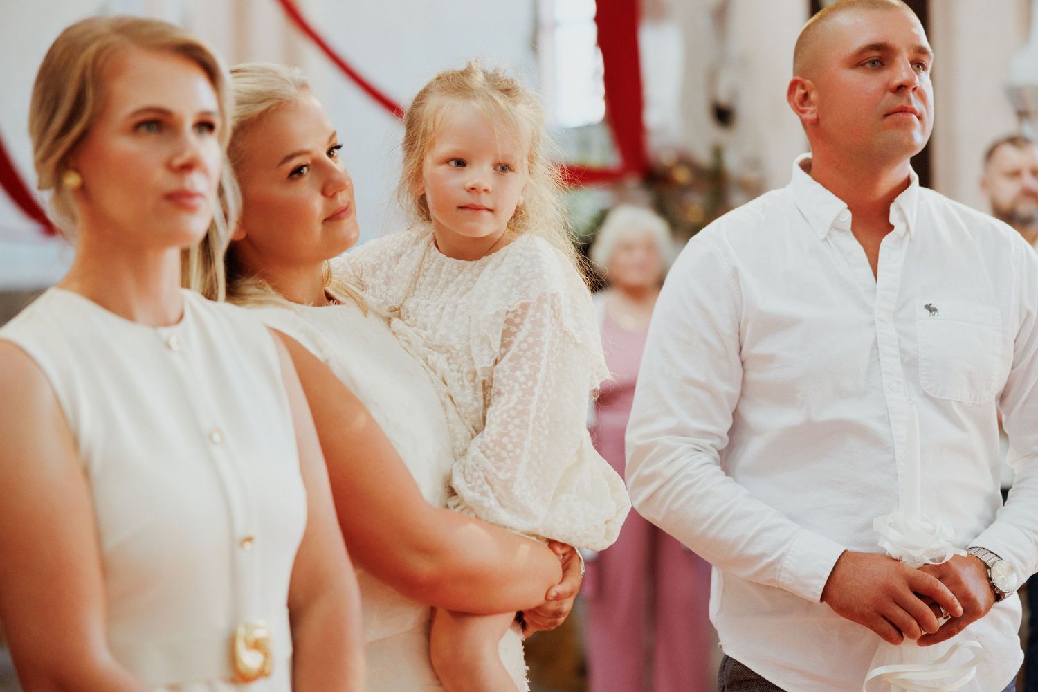 A family in white attire stands together during a formal celebration with red and white decor in the background.