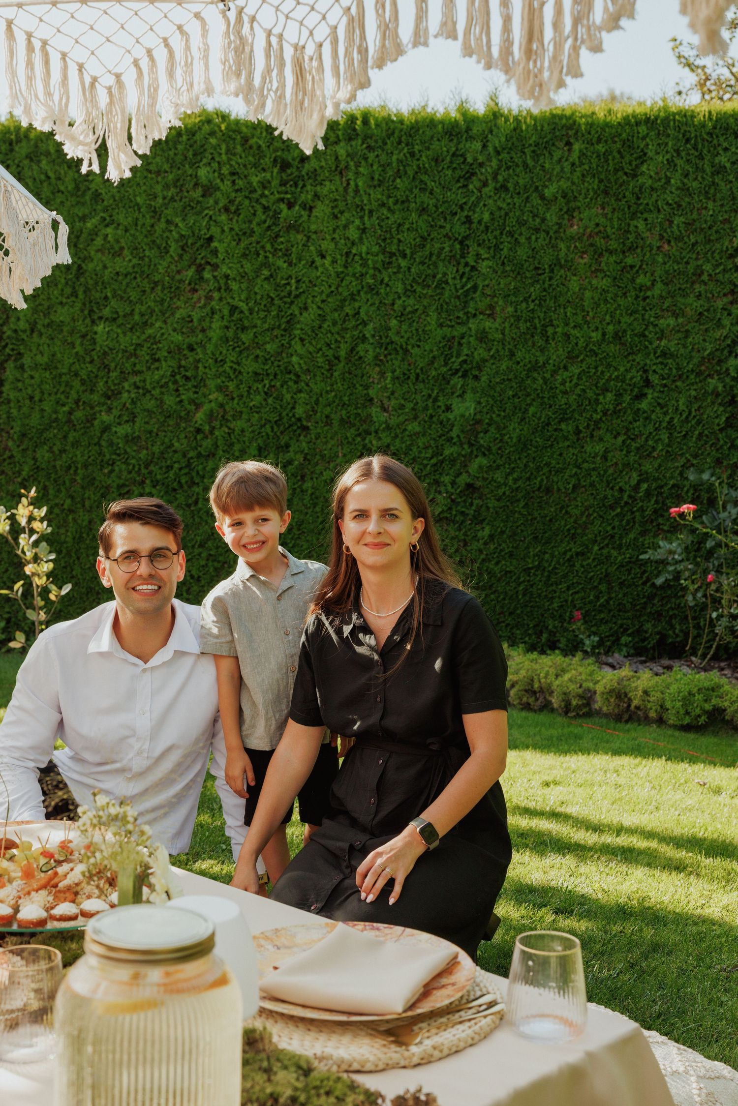 Family gathering around outdoor table with macrame decor and green hedge backdrop.