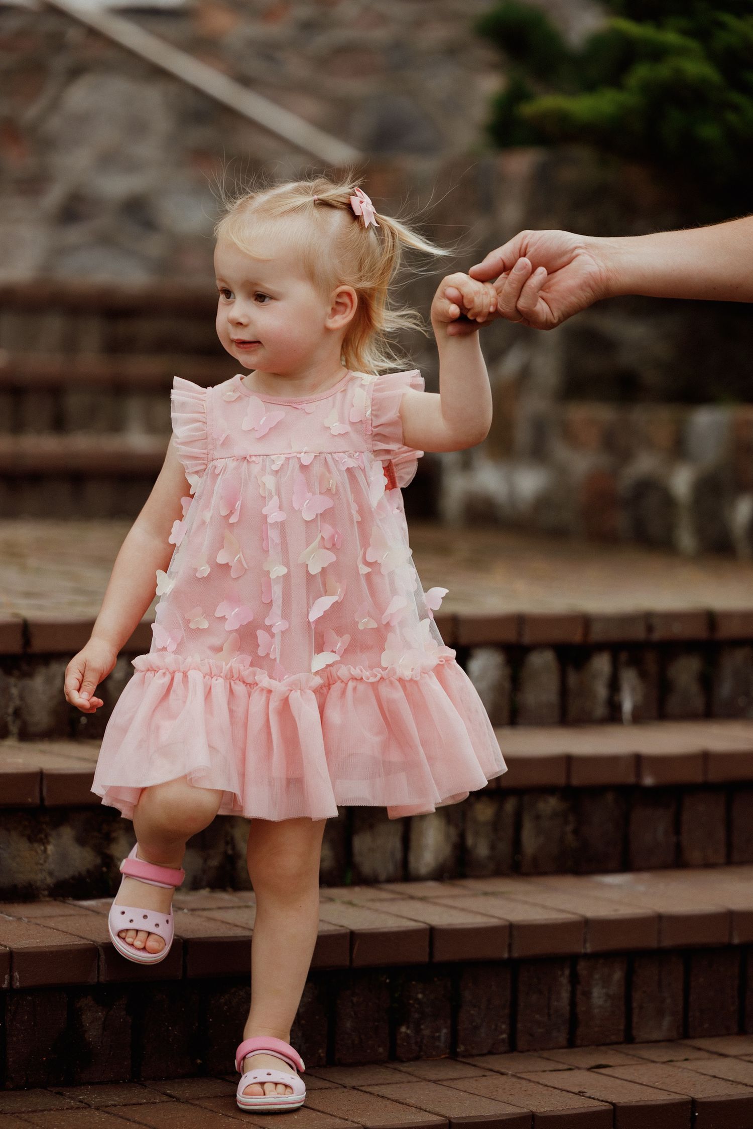 Toddler in pink ruffled dress walks up brick steps while holding an adult's hand.