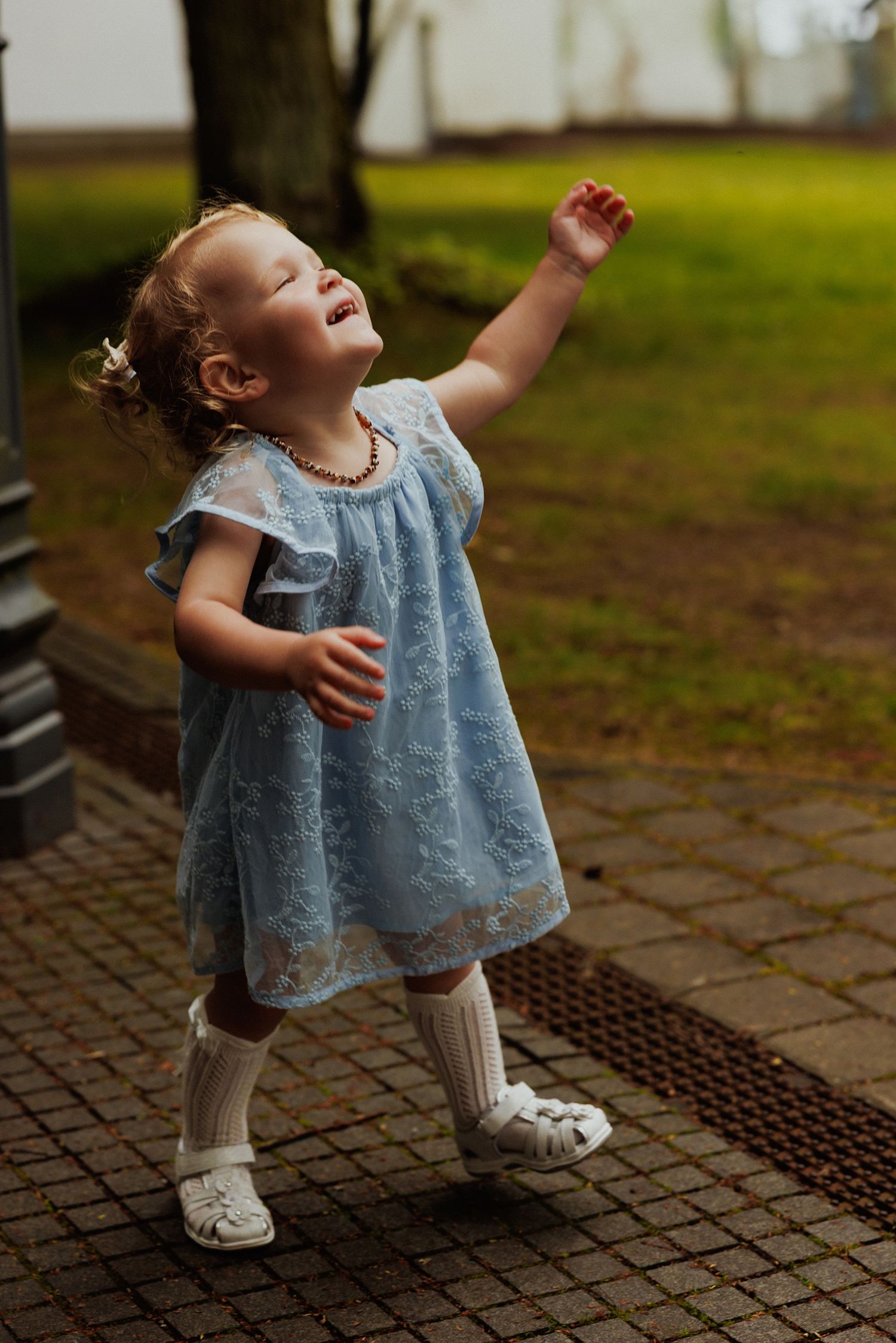 A small child in a blue dress and white shoes looks upward while standing on a brick paved surface outdoors.
