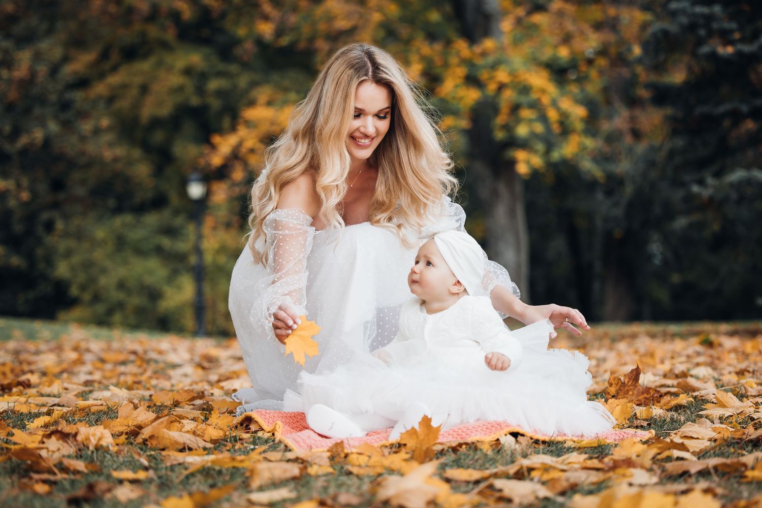 A person in a white dress sits joyfully among fallen autumn leaves in a park setting during fall season.