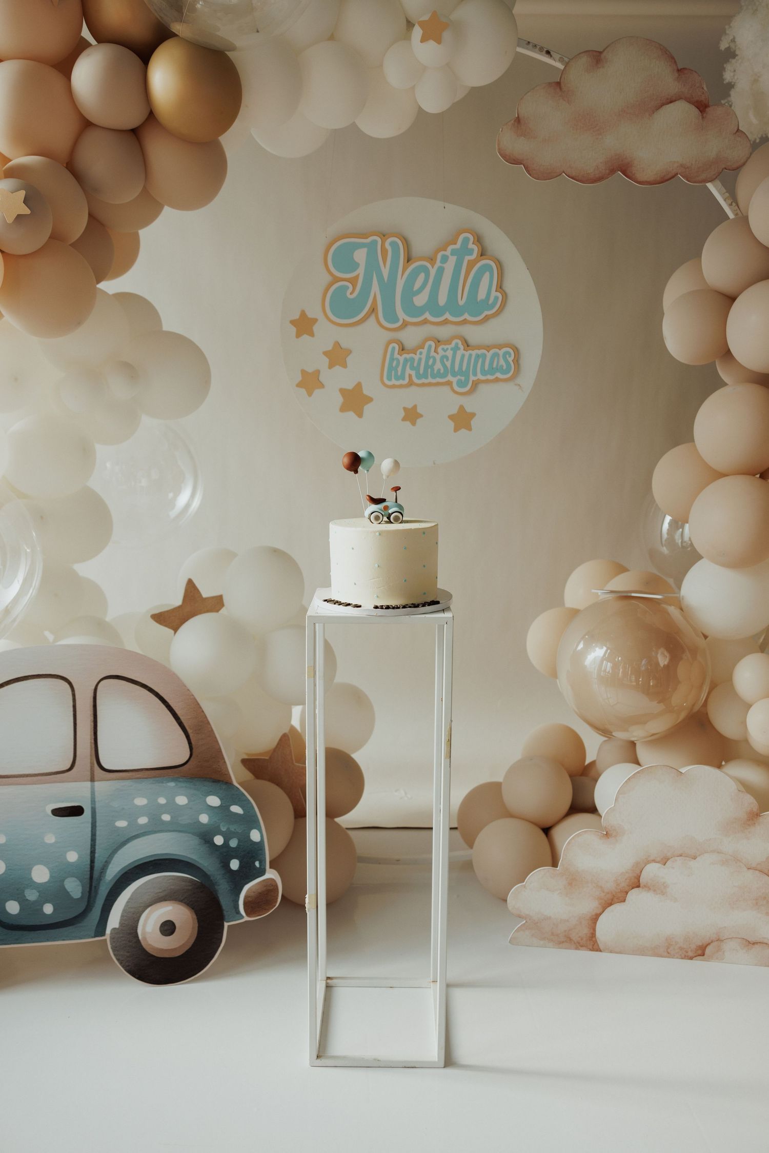 Baby shower backdrop with beige balloon arch and decorative vintage vehicle cutouts.