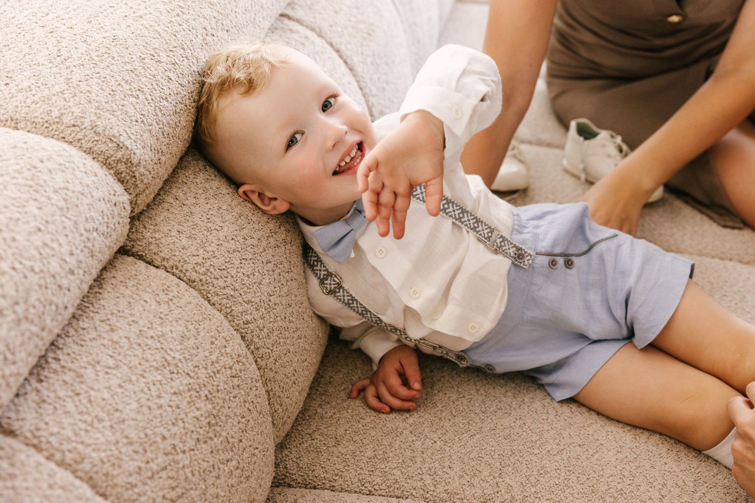 Baby in white shirt and suspenders lying on beige couch.