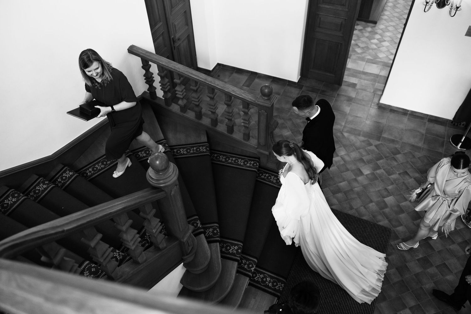 A wedding party descends a grand wooden staircase in black and white photography.