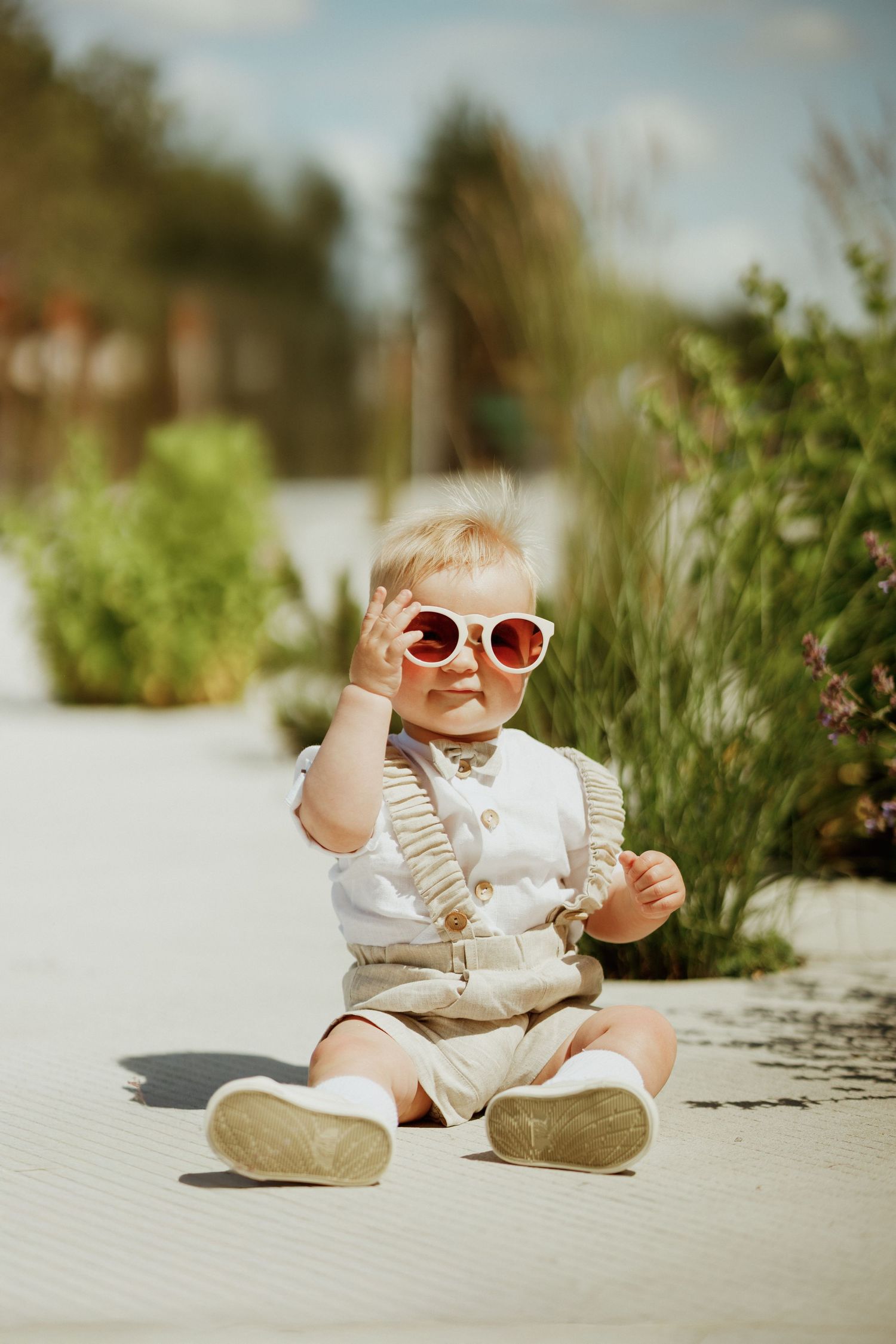 Baby in white and beige outfit wearing sunglasses while sitting outdoors on white surface.