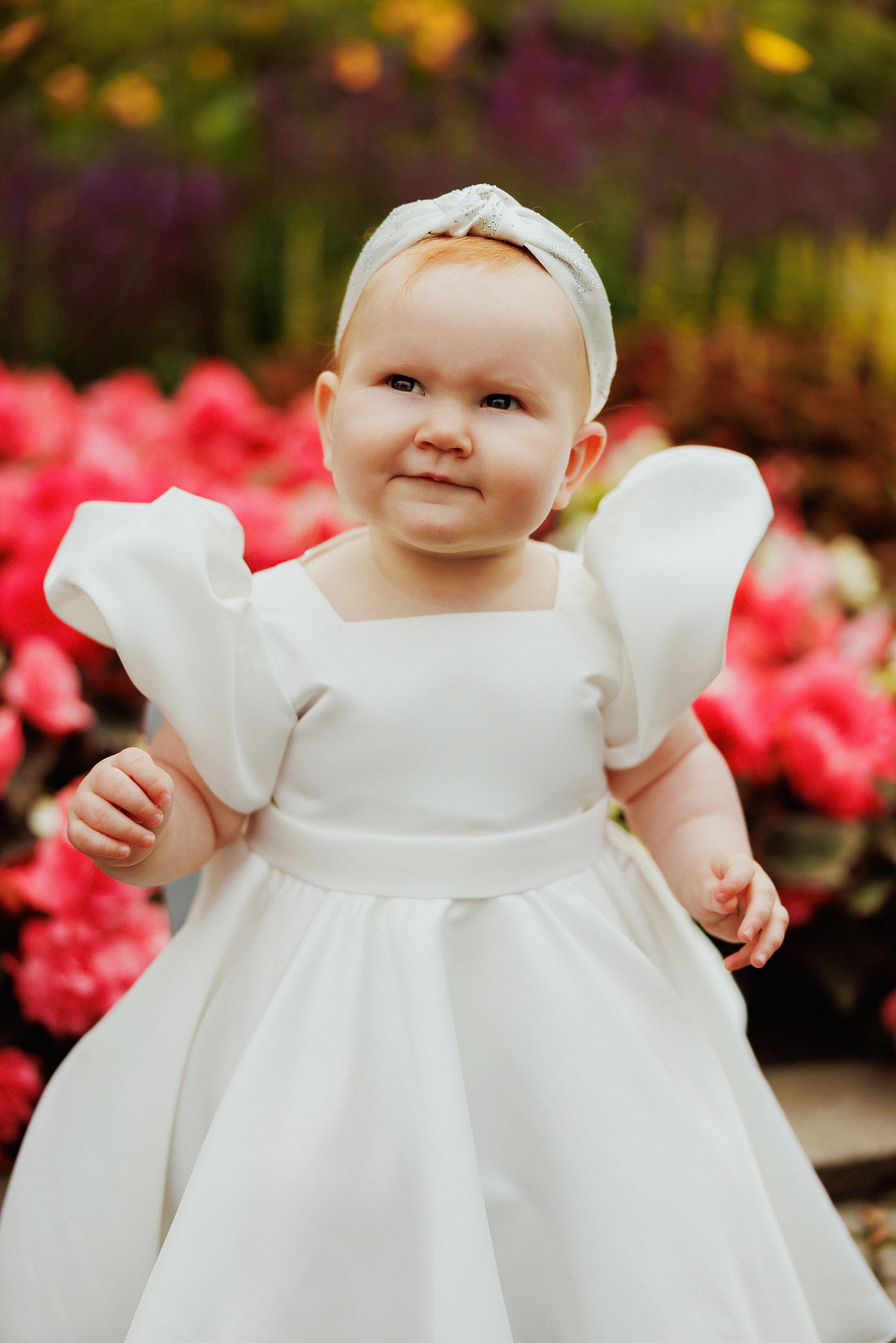 Baby in a flowing white christening gown sitting among colorful garden flowers.