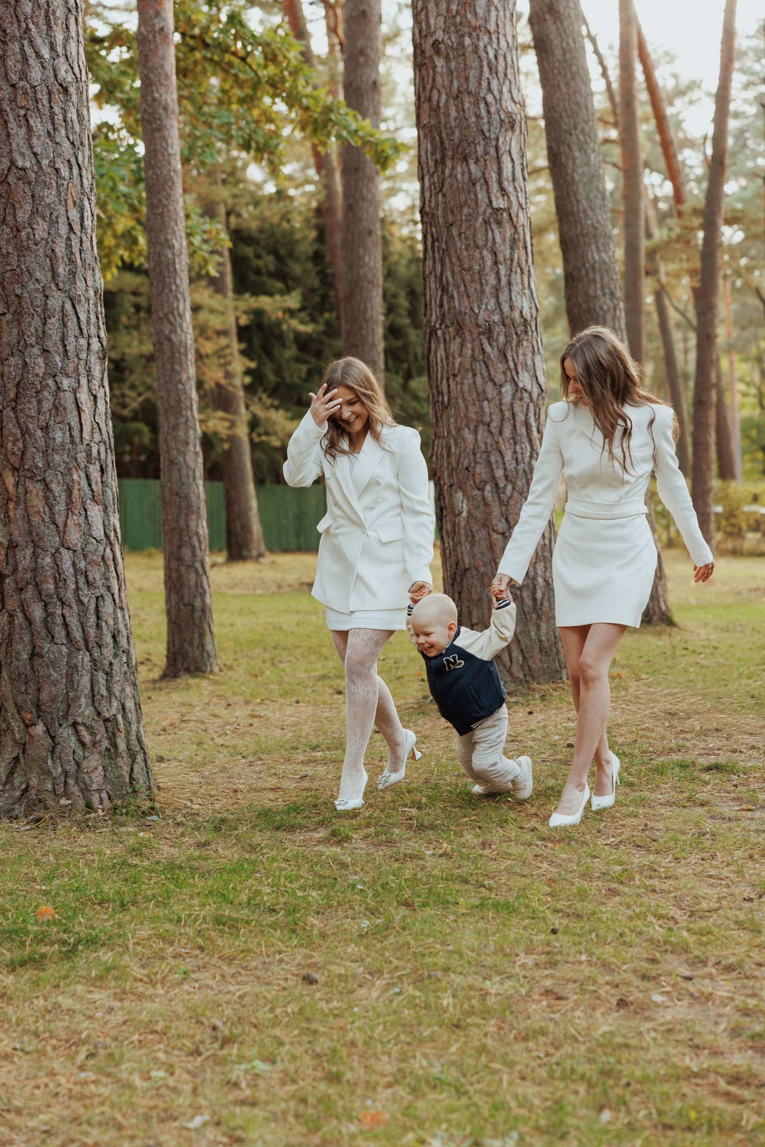A playful moment in a pine forest as people in white outfits swing a small child between them.