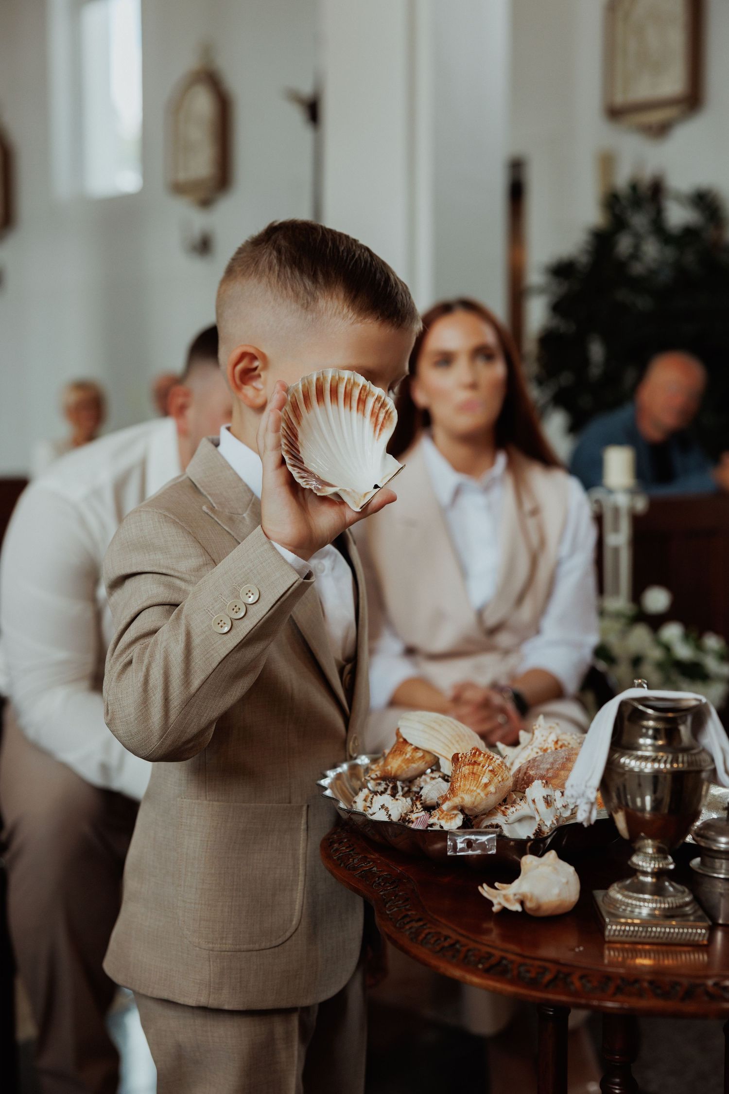 A young attendee in a beige suit holds a seashell to their face during an elegant indoor gathering.