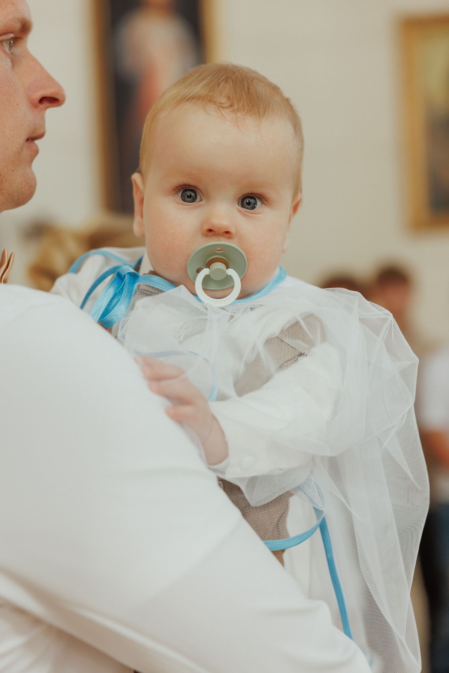 Baby in white christening gown with blue sash and pacifier during baptism ceremony.
