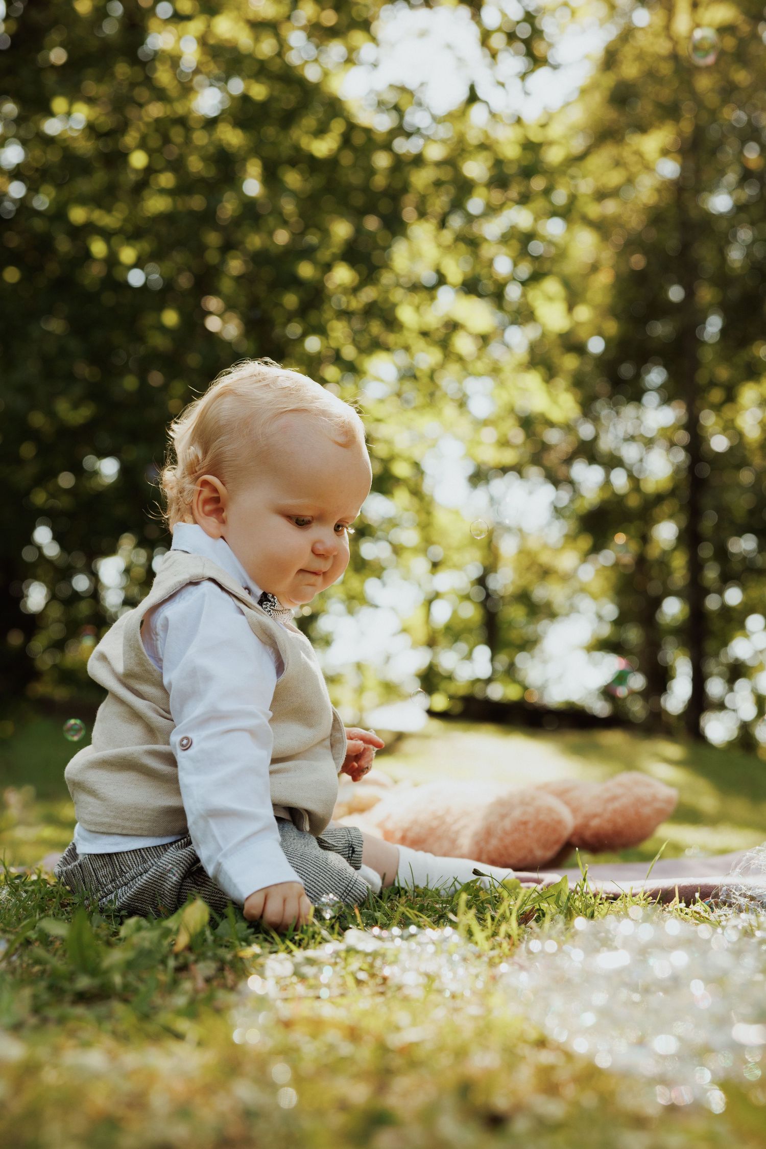 Baby in white clothing sitting on grass in a sunny garden with bokeh lighting effects.