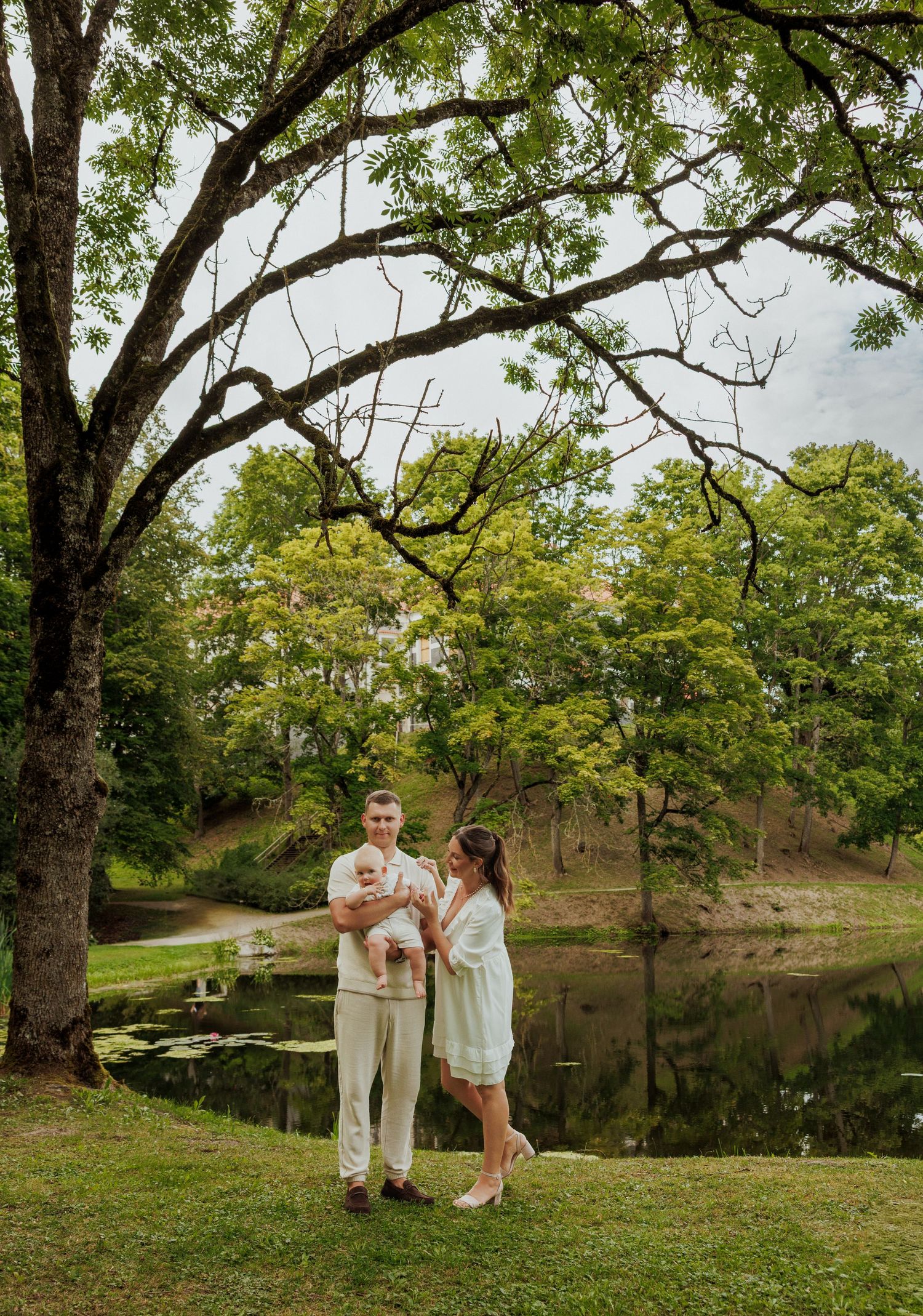 Couple shares a tender moment under large trees beside a tranquil pond.