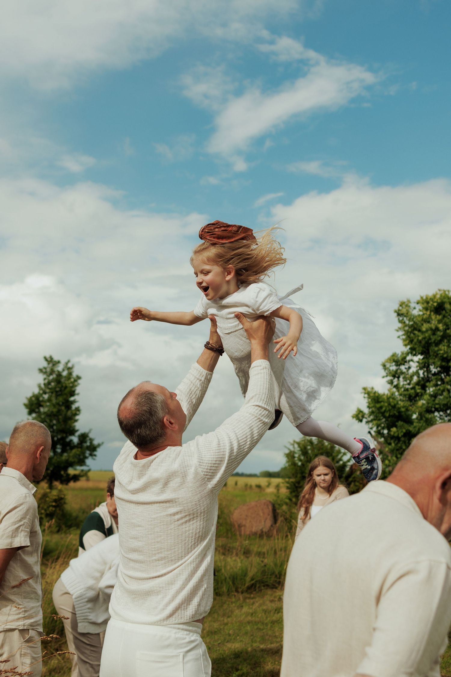 Man in white lifting a laughing girl in a white dress outdoors on a sunny day in a green field.