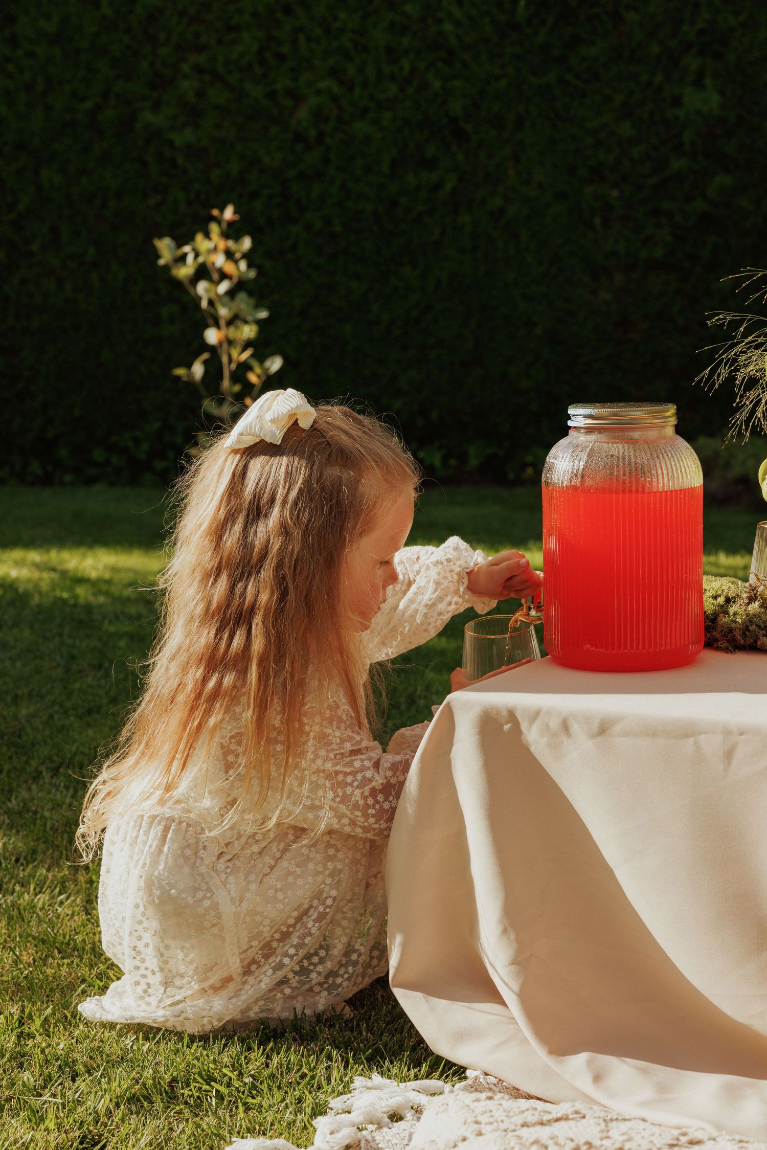 A young girl in lace dress pours a drink from a large glass dispenser at an outdoor party.