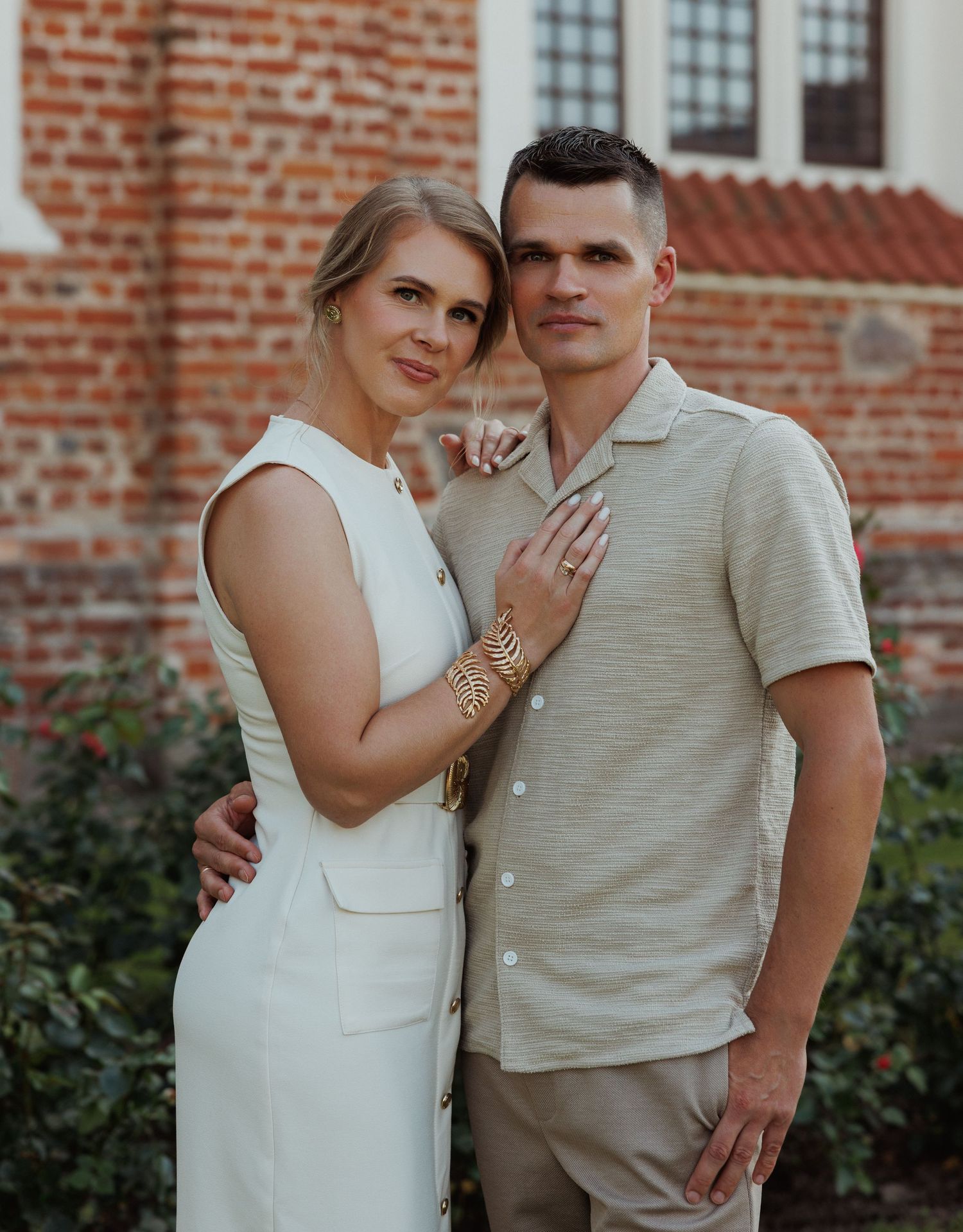 Couple in coordinating white and beige outfits embrace against brick building backdrop.