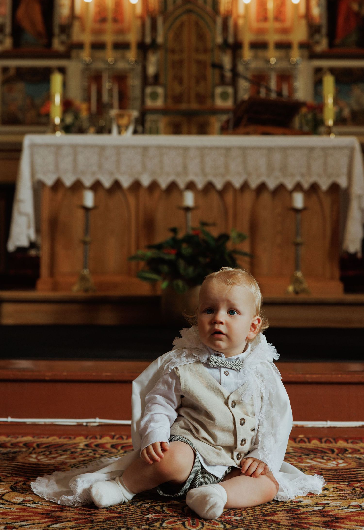 Two infants in white christening outfits sit on an ornate red carpet in front of a church altar.