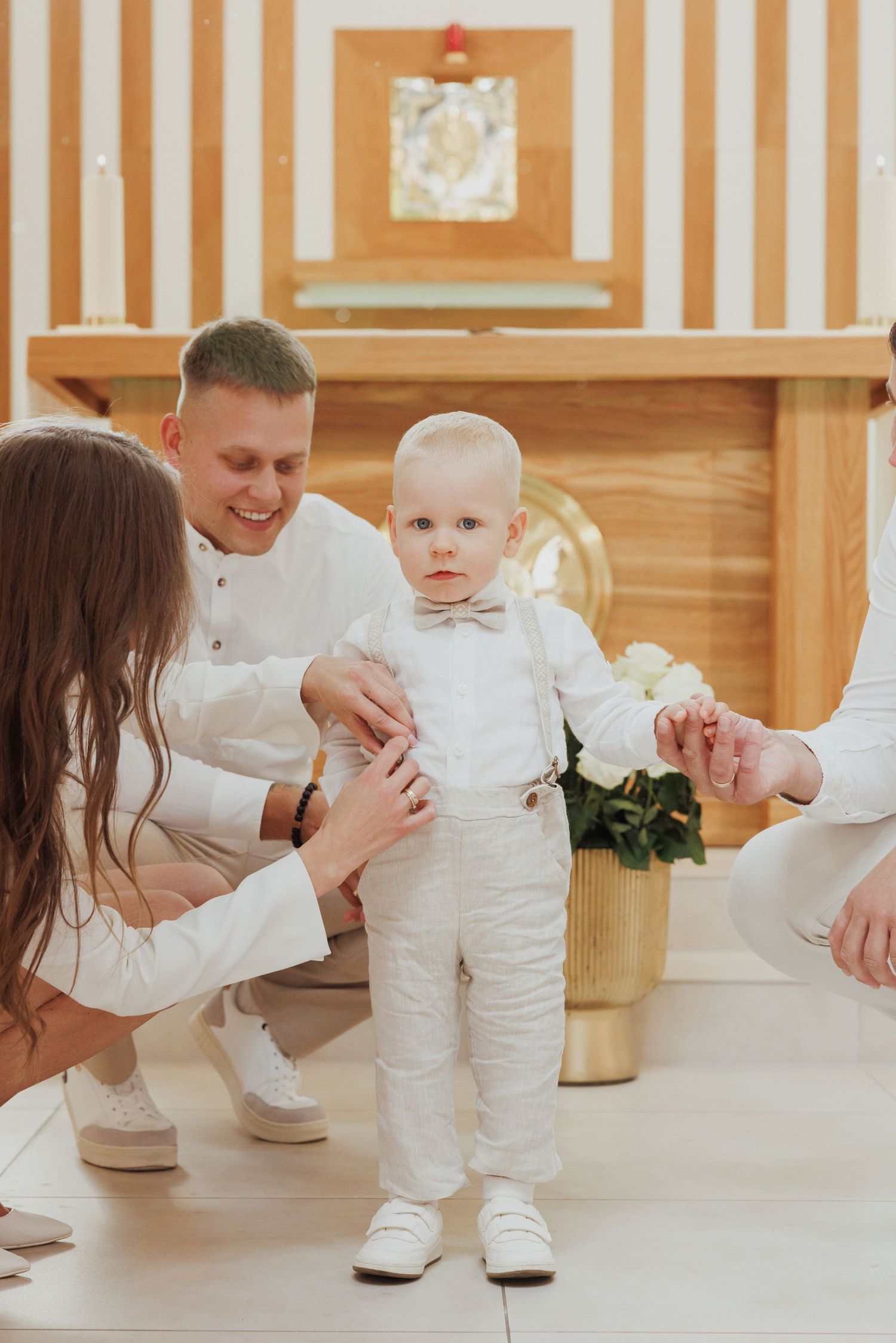 A toddler in all white stands between adults during a special celebration in a room with striped walls.