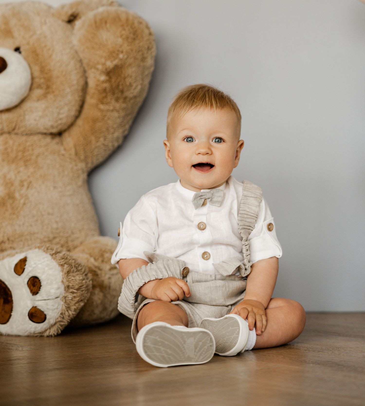 Baby in white and beige outfit sitting on wooden surface with teddy bear in background.