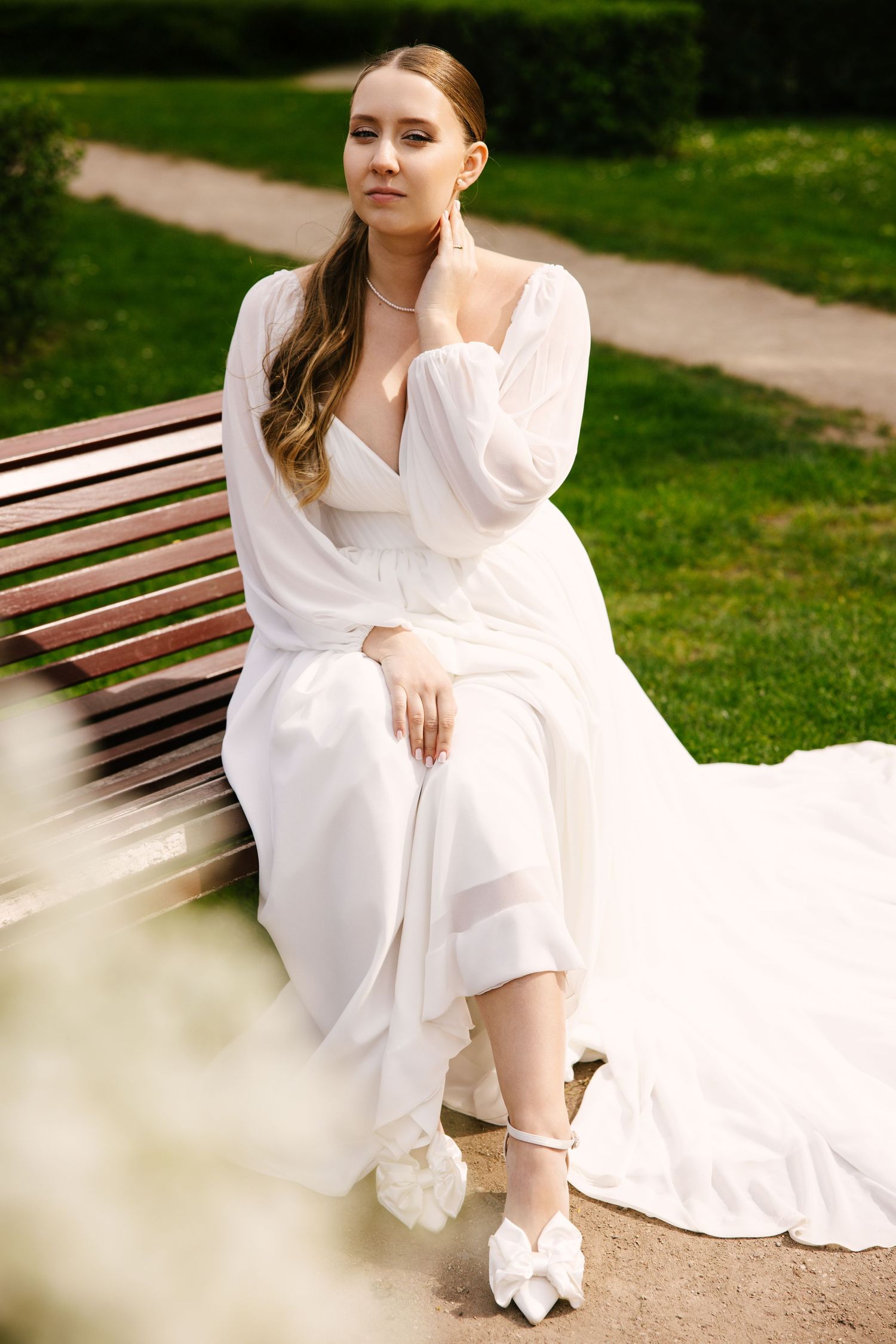 A bride in a flowing white dress sits elegantly on a park bench with delicate bridal shoes.