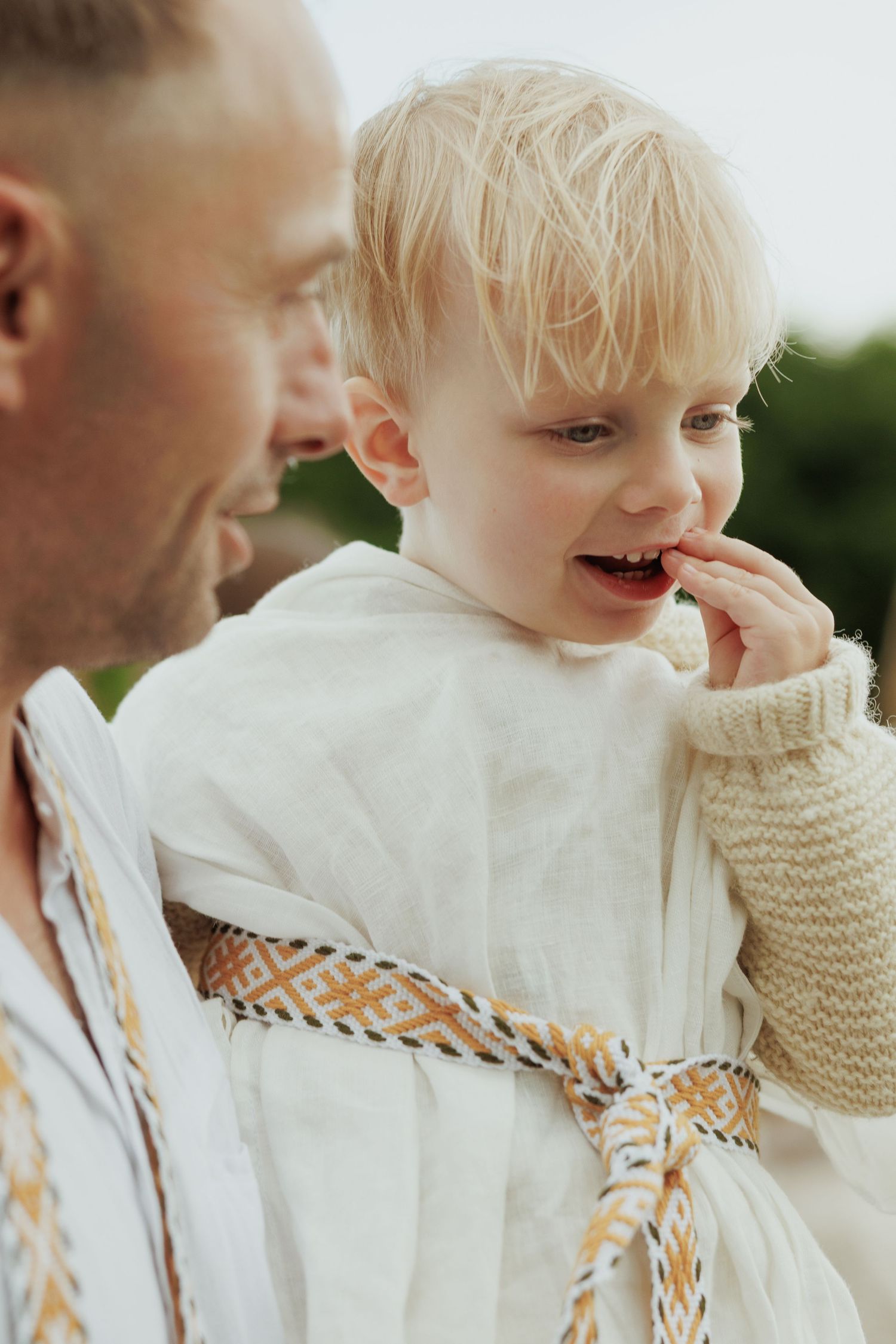 Blonde toddler in traditional embroidered white linen outfit eating a berry while being held by a man outdoors.