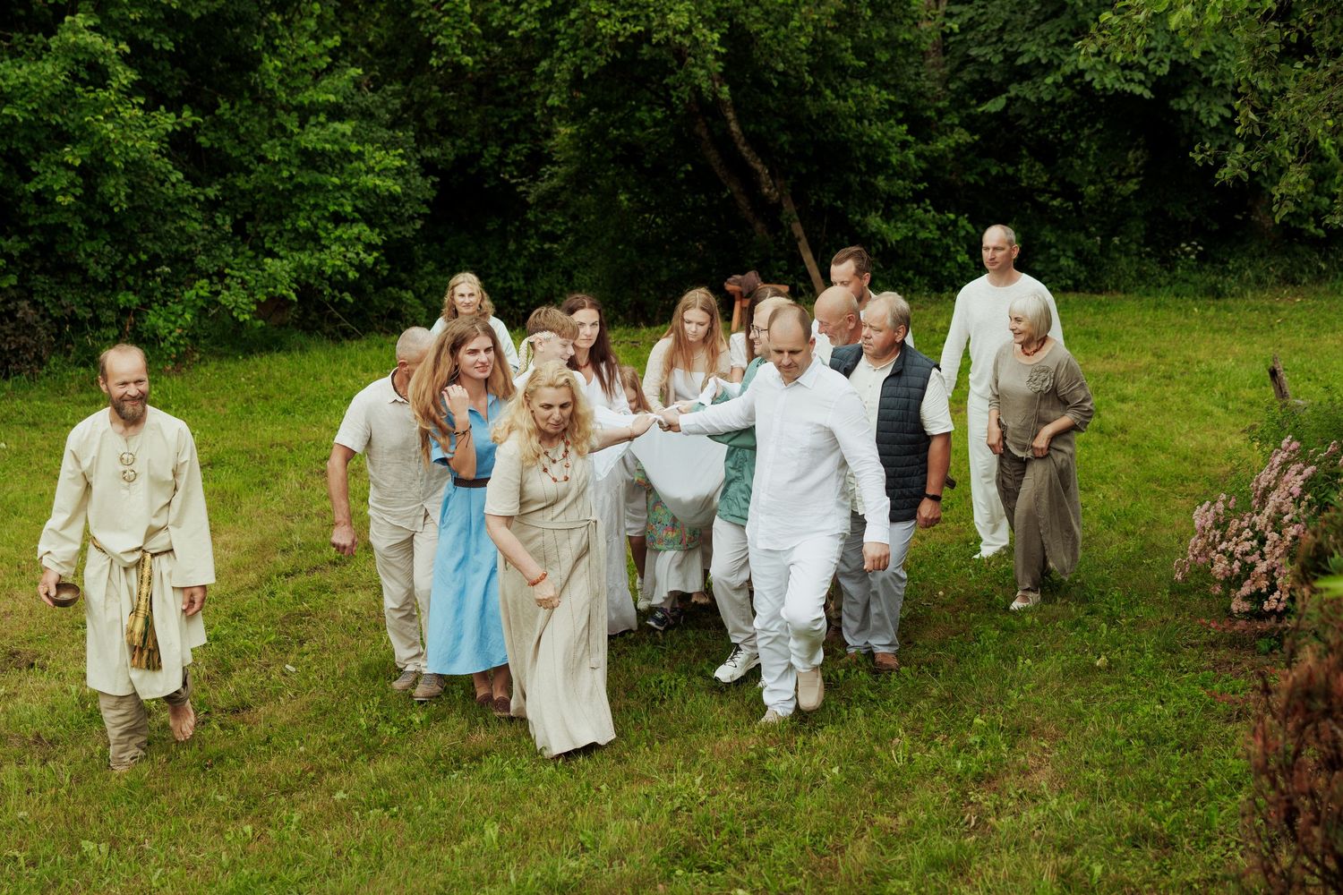 Group of people dressed in white and linen clothing walking together on a green grassy field surrounded by trees.