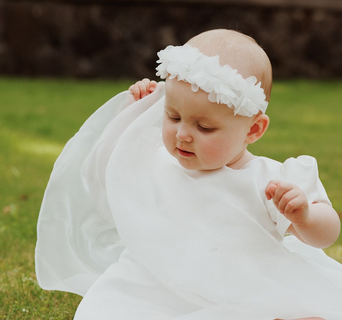 A baby in a flowing white christening gown crawls on green grass during a sunny outdoor photo session.