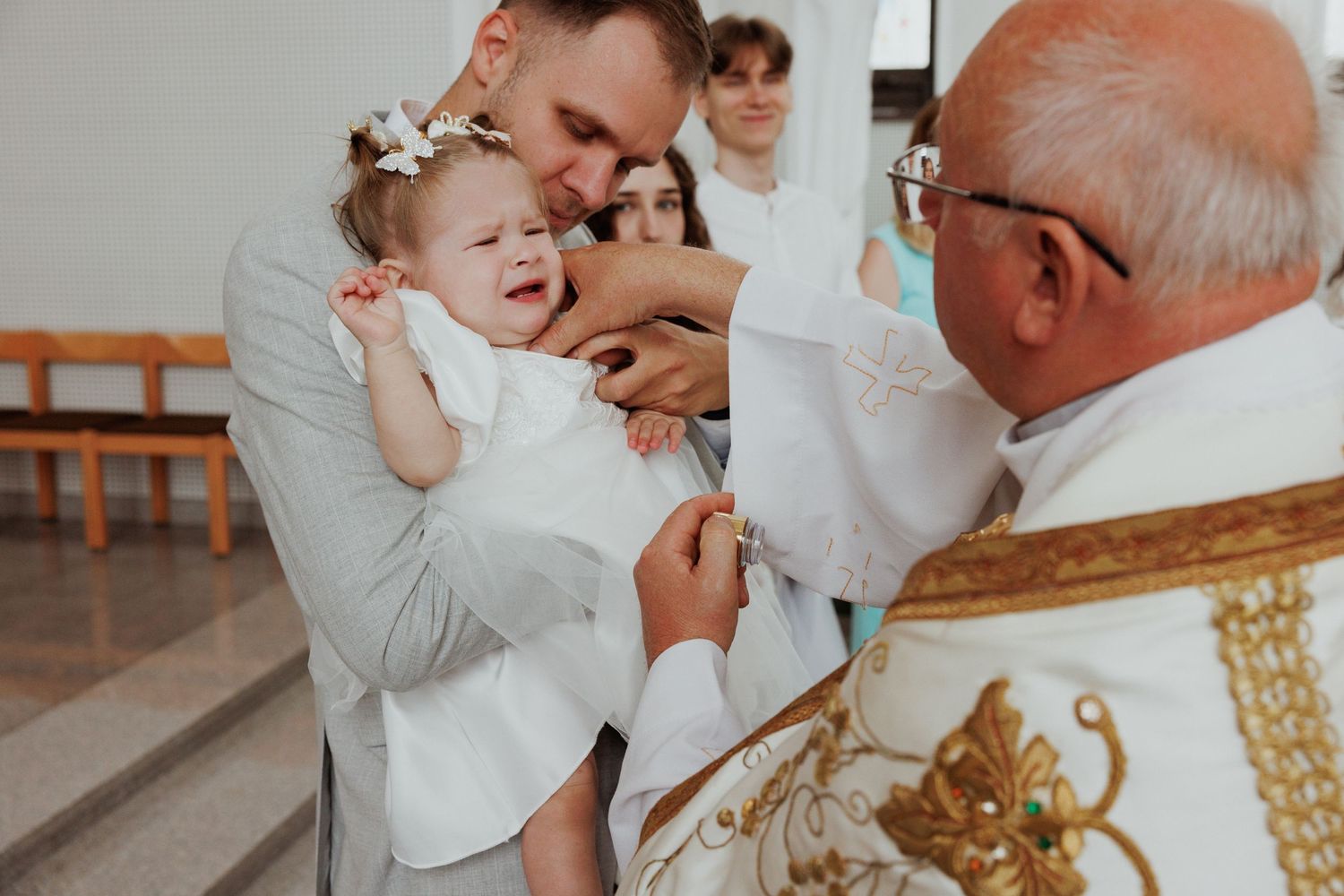 A tender moment captured during a baptism ceremony as a baby in white is held over an ornate church baptismal font.