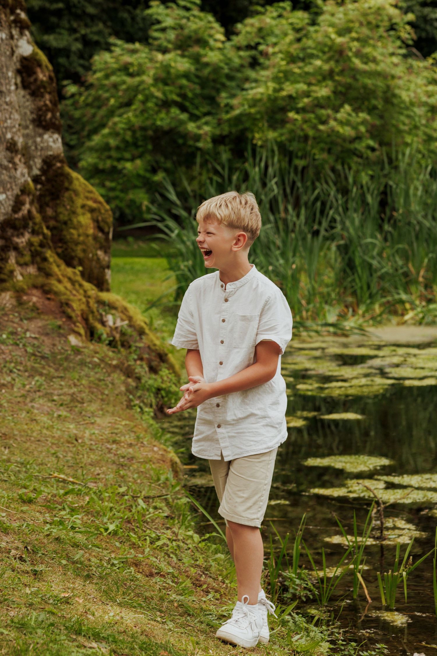 Young child in white plays near a pond with lily pads on a sunny day.