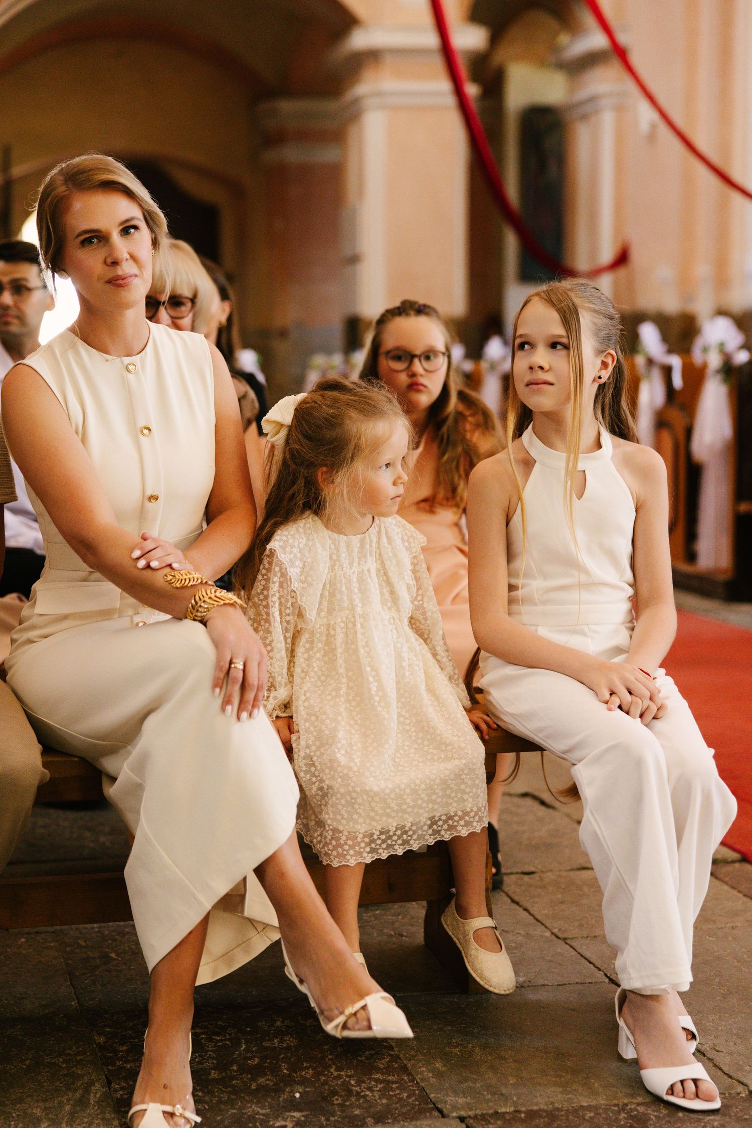 Family members dressed in white formal attire sit together at an elegant indoor venue with red carpet and arches.