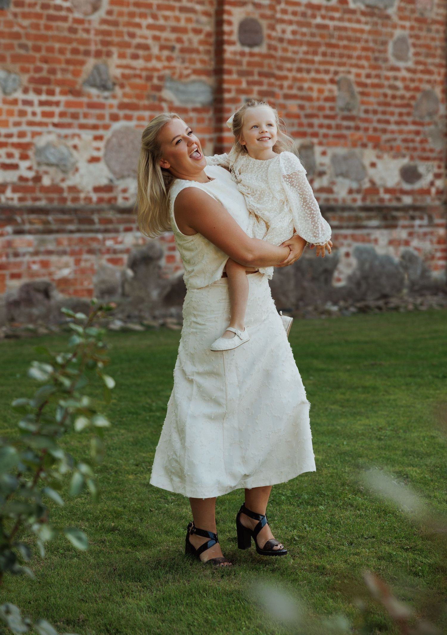 Playful interaction between two people in white dresses against a brick wall backdrop.