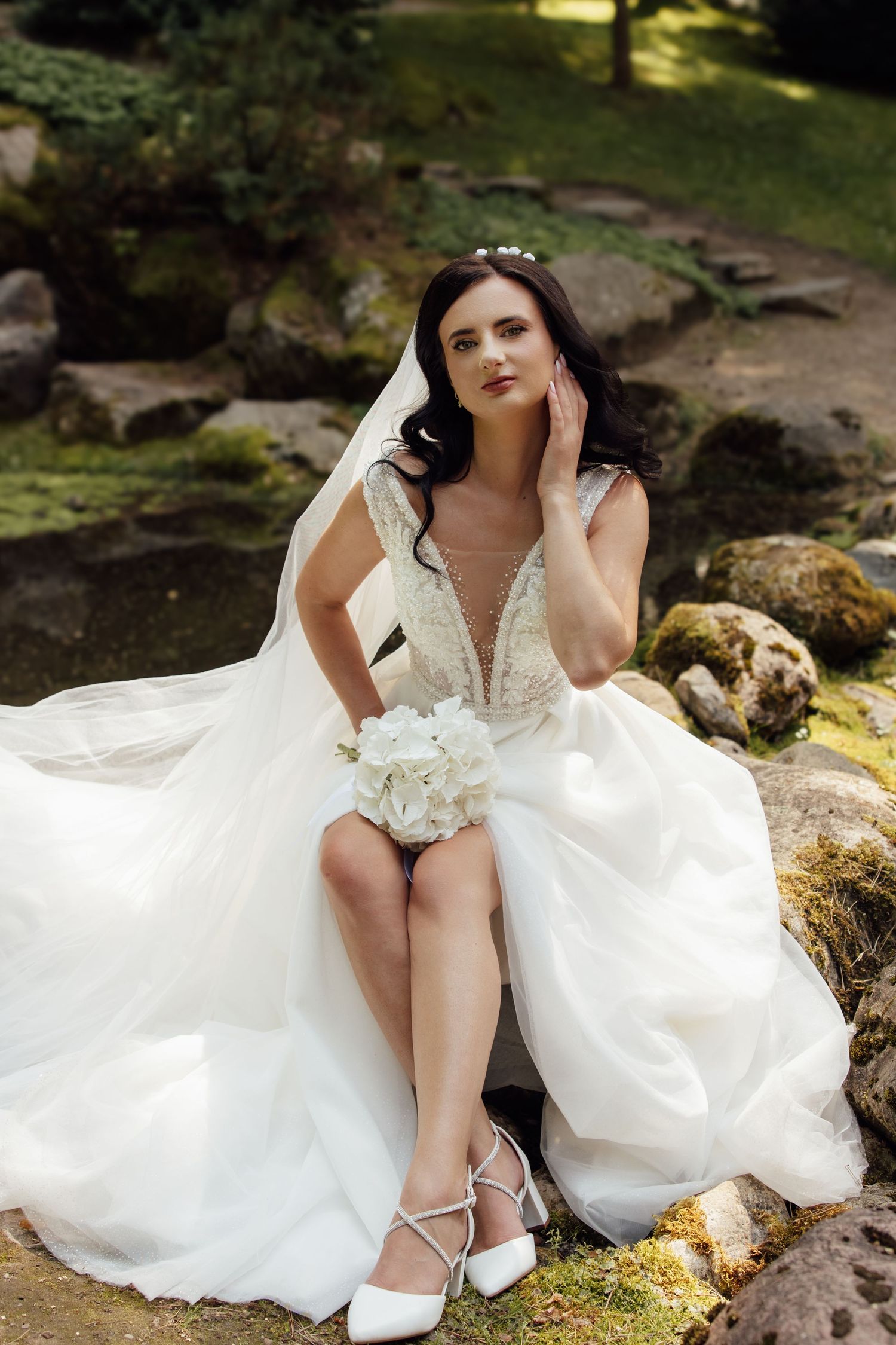 Elegant bride in white lace gown sits on mossy rocks in garden, holding hydrangea bouquet with veil flowing.