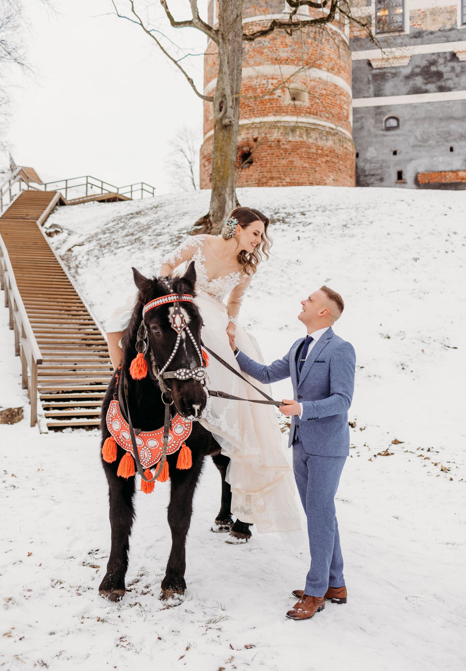 Winter wedding scene with couple and decorated horse in snow against brick building with stairs.