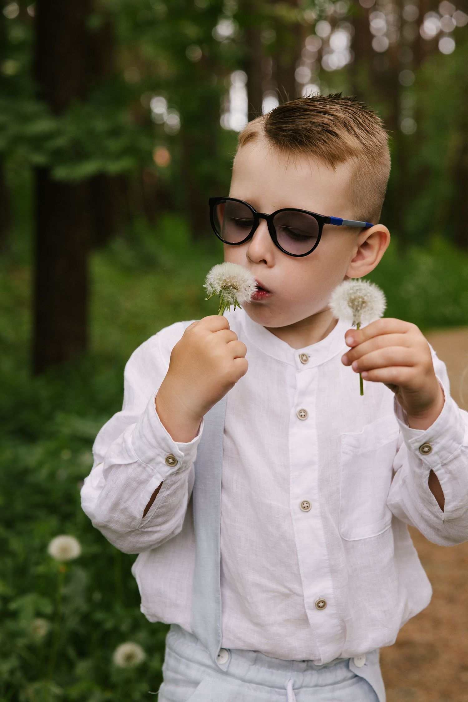 Child in white shirt and glasses blowing dandelion seeds in a forest setting.