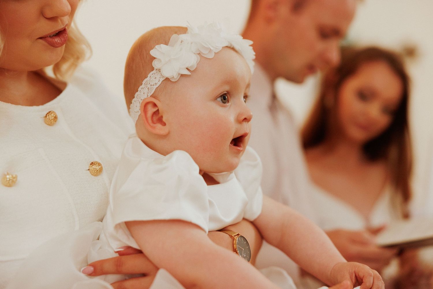 A baby in white clothing reaches out while sitting on a wooden bench with decorative flowers in the background.