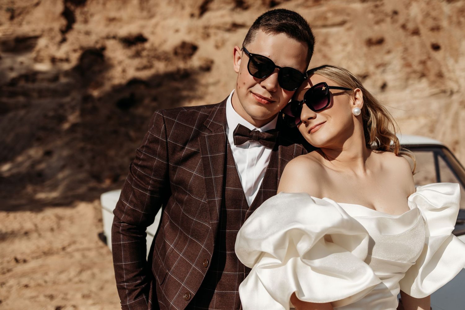 Elegant couple in formal wedding attire pose together against rocky desert backdrop wearing stylish sunglasses.