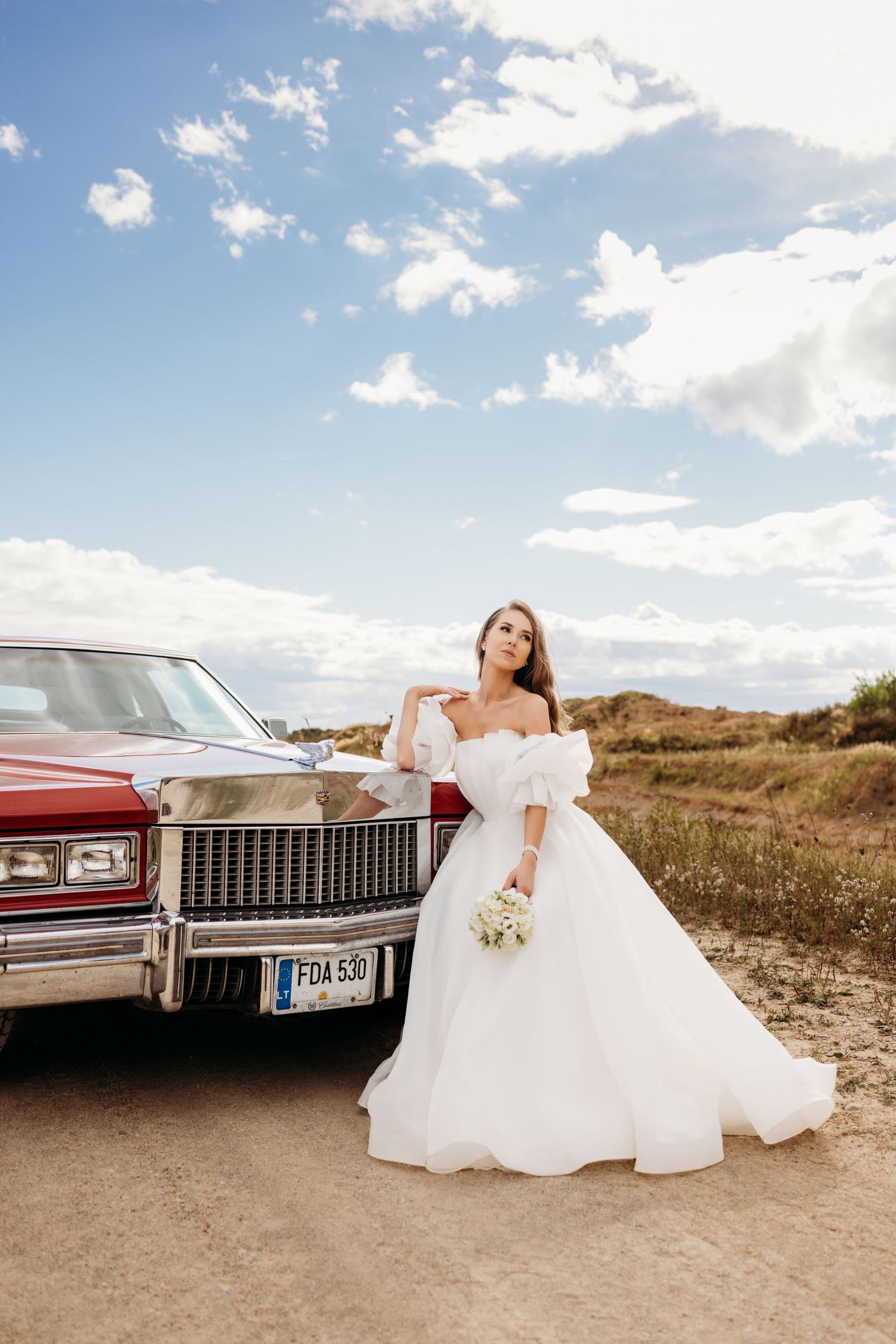 Bride in off-shoulder white gown poses beside classic red Cadillac under blue sky.