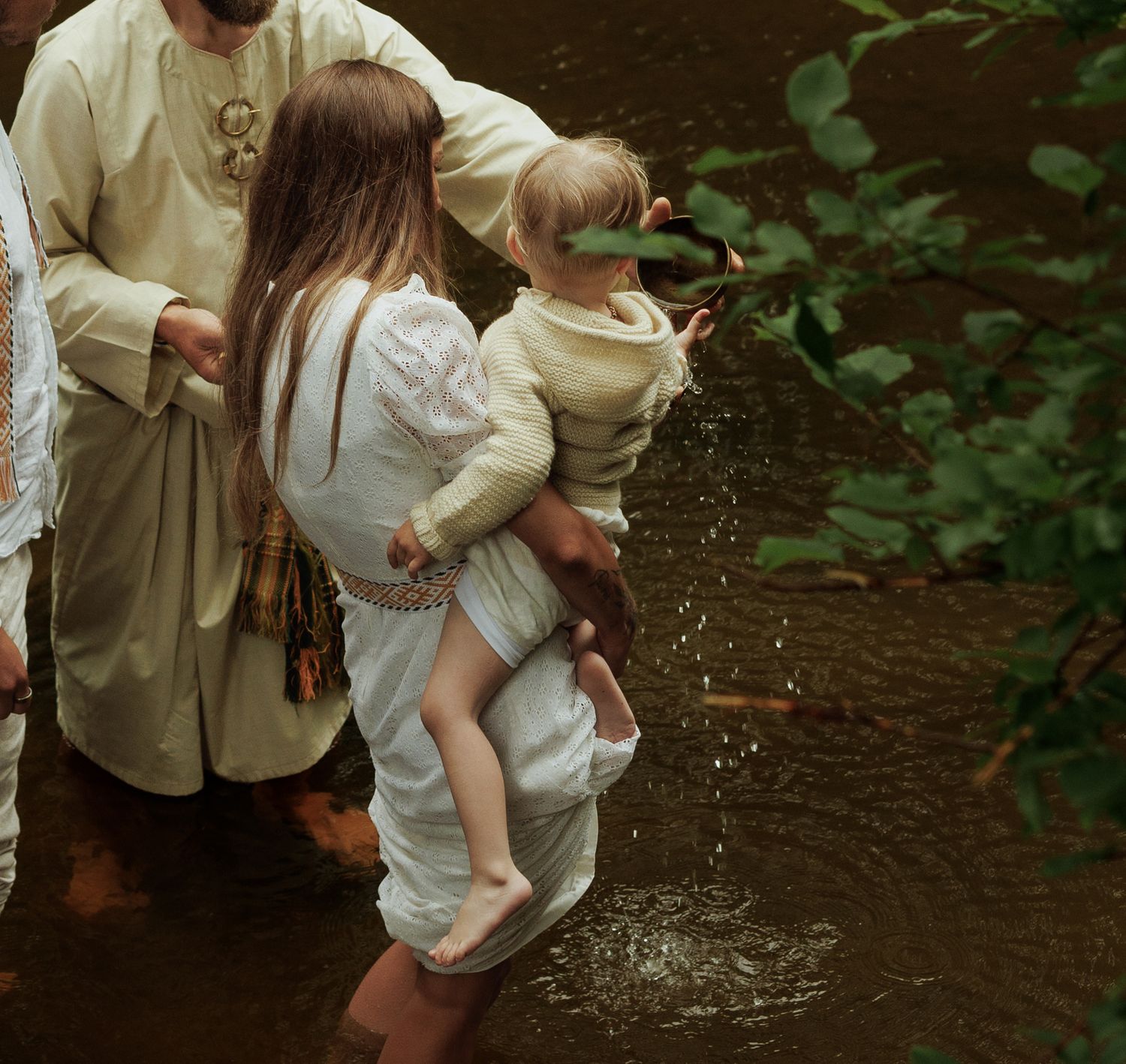 Woman in white dress holds toddler over a creek, with a robed figure nearby, in a lush green outdoor setting.
